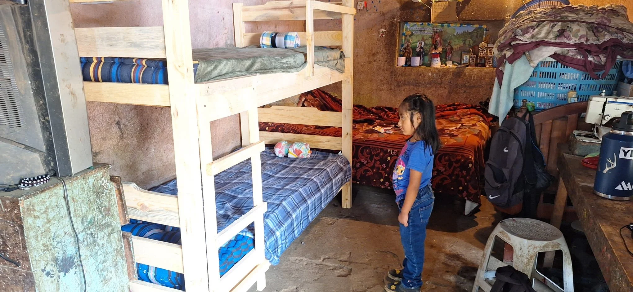 A young girl standing in a rustic room with bunk beds, a bed with a blanket, and various items scattered around including a backpack, a small stool, and decorative figures on the wall.