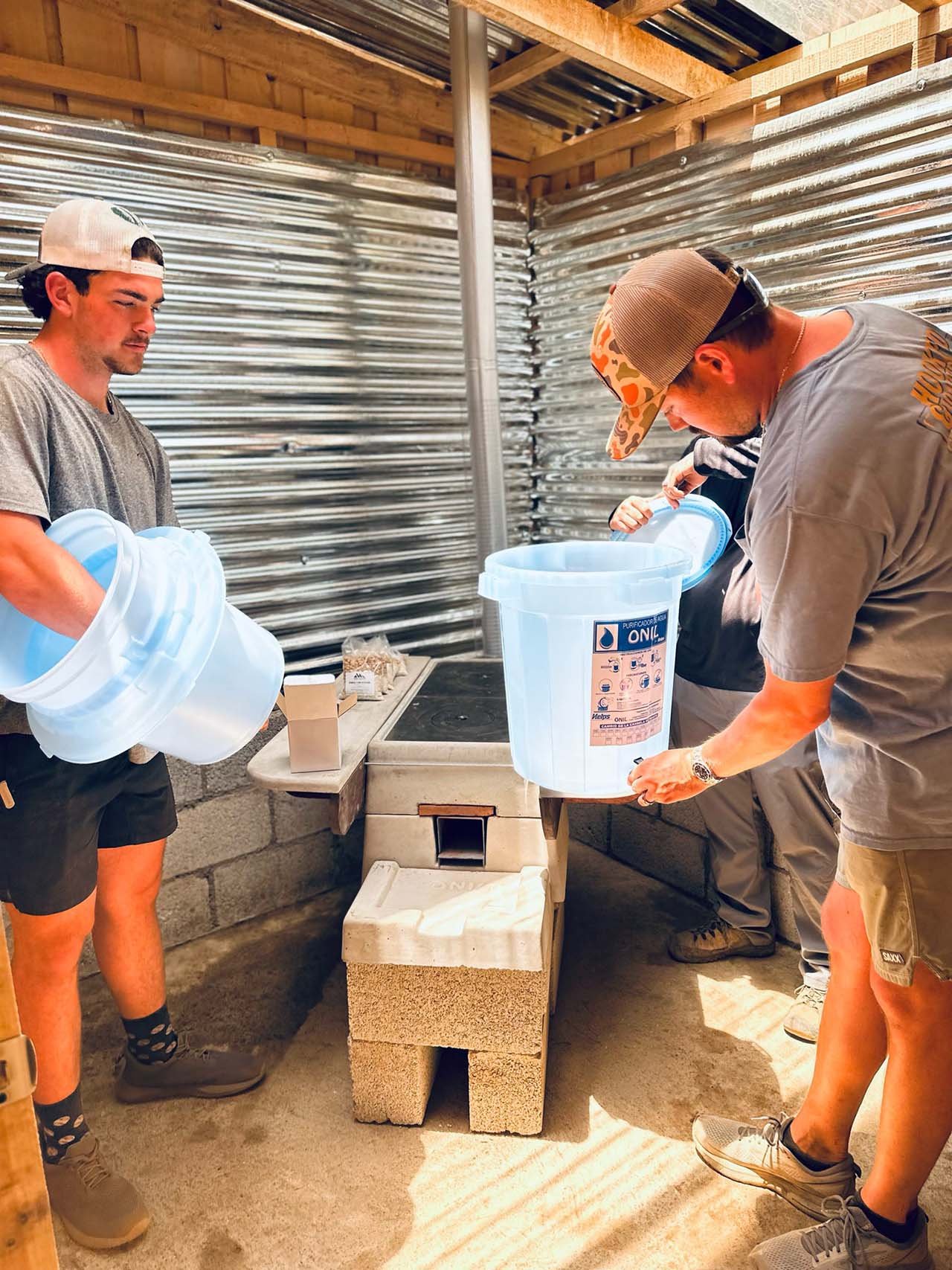 Two young men working together in a construction or workshop setting, pouring or handling large plastic containers, with a metal and wood structure in the background.