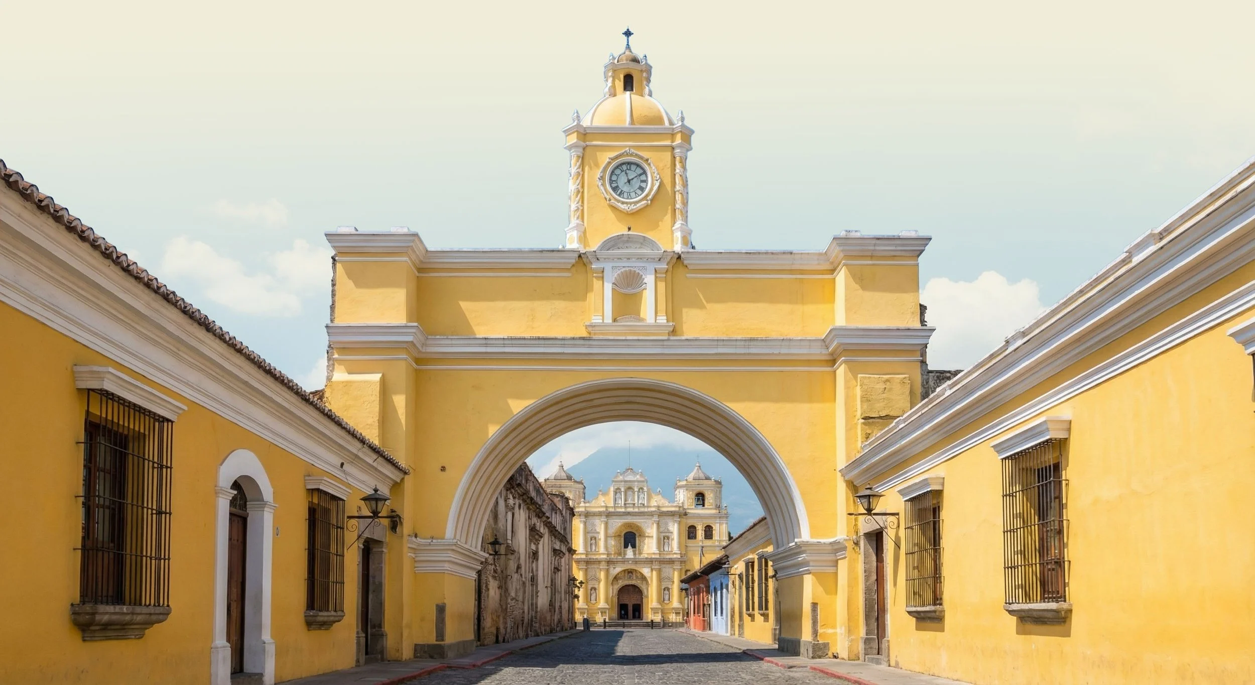Yellow historic building with arch and clock tower, leading to a church in the distance, cobblestone street