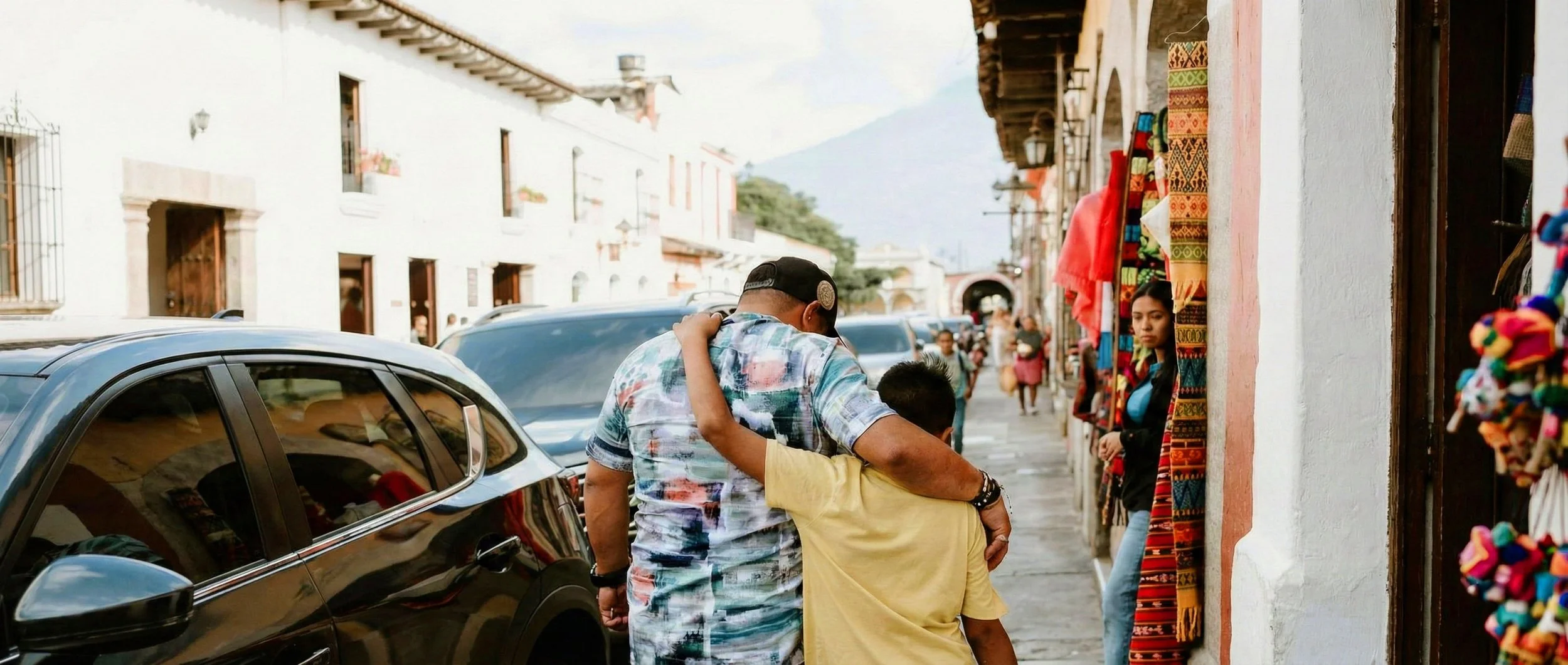 A man and boy hugging on a bustling street with parked cars, pedestrians, colorful textiles, and white colonial-style buildings with flower boxes, set against a mountain backdrop.
