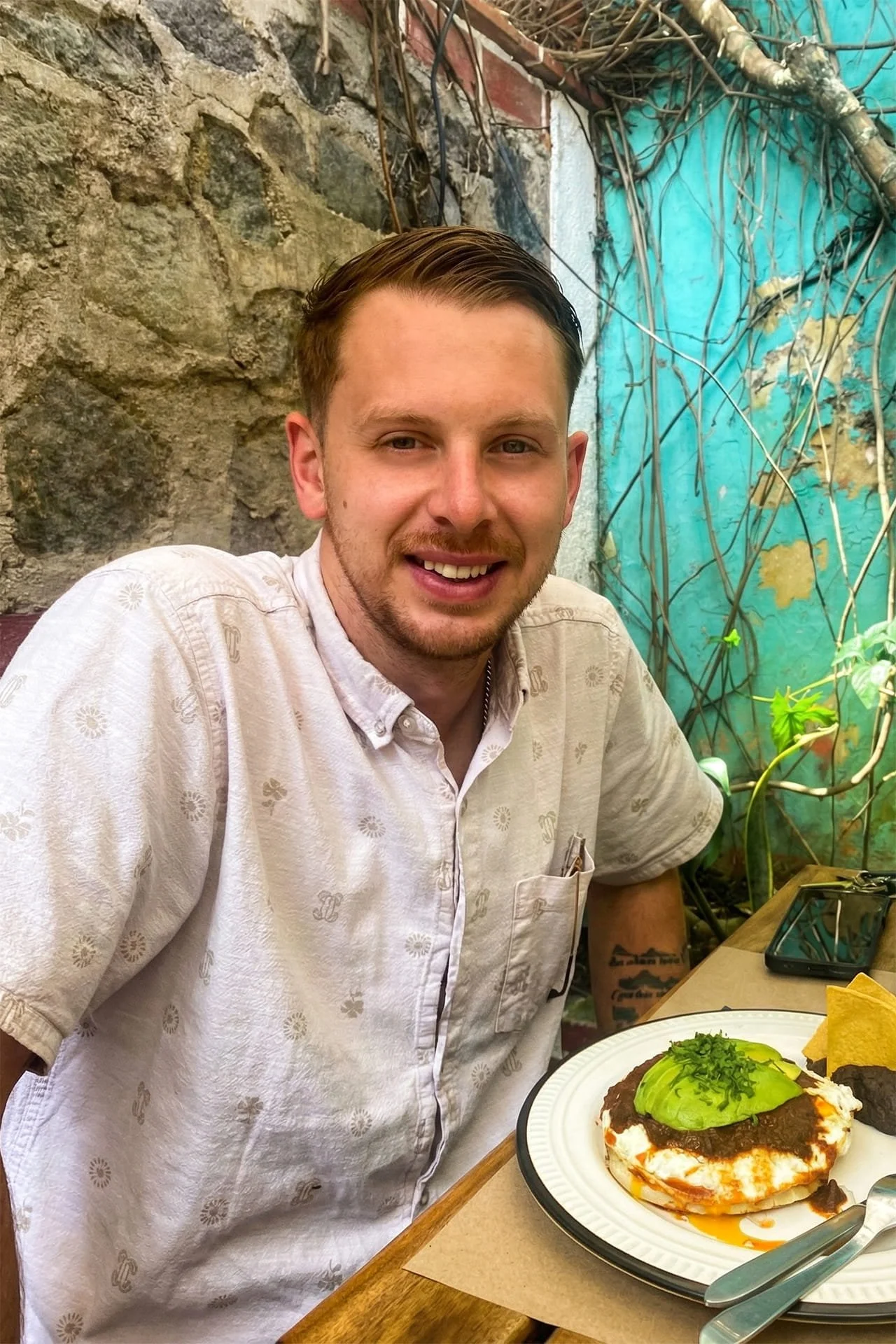 A man smiling at a table with Mexican food including an avocado-topped dish, black beans, and fried plantains, with a stone wall and vine-covered turquoise wall in the background.