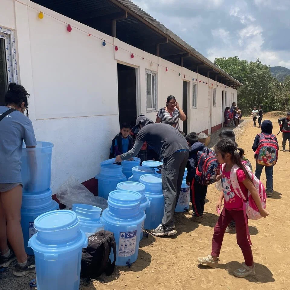 Children and adults outside a building with string lights, receiving bottled water from large blue containers on a dirt ground.