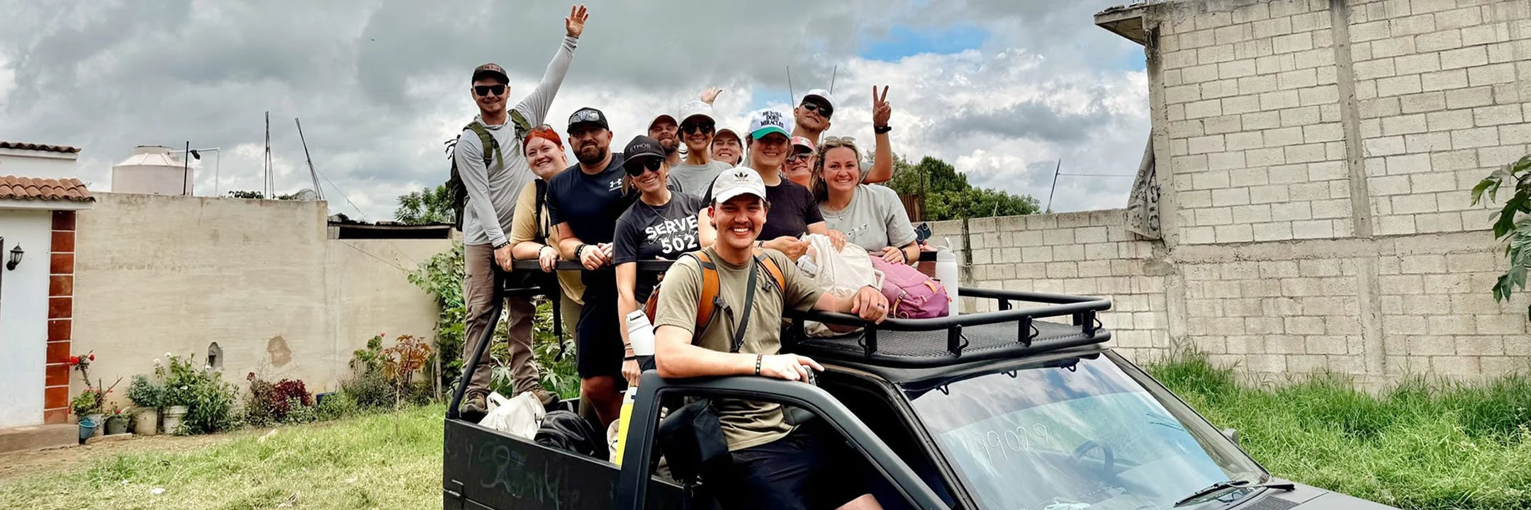 A group of people on a small black vehicle, smiling and making peace signs, outdoors on a cloudy day with a brick wall and plants in the background.