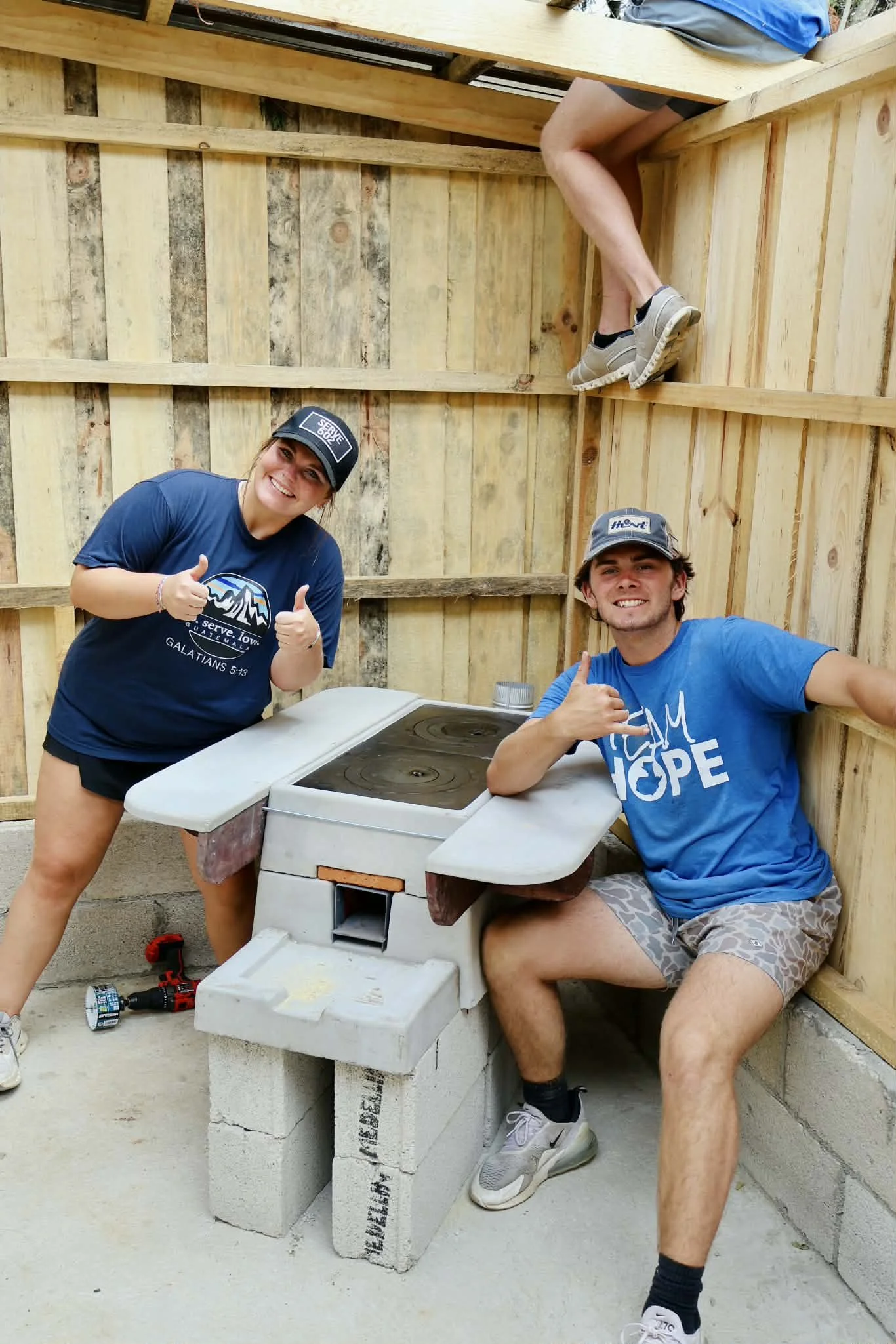 Two young people in a partially built outdoor structure, smiling with thumbs up, around a makeshift stove made of concrete blocks and a round metal top, one woman standing and a man sitting, with a wooden fence in the background.