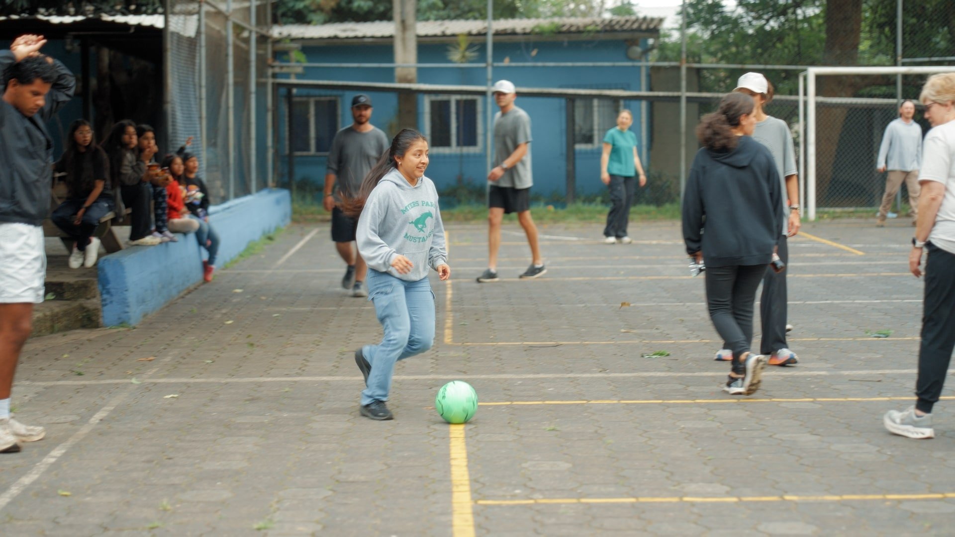 A girl is playing soccer on a court with a green ball. Several people are present, some watching from benches, others standing and walking, with trees and a blue building in the background.