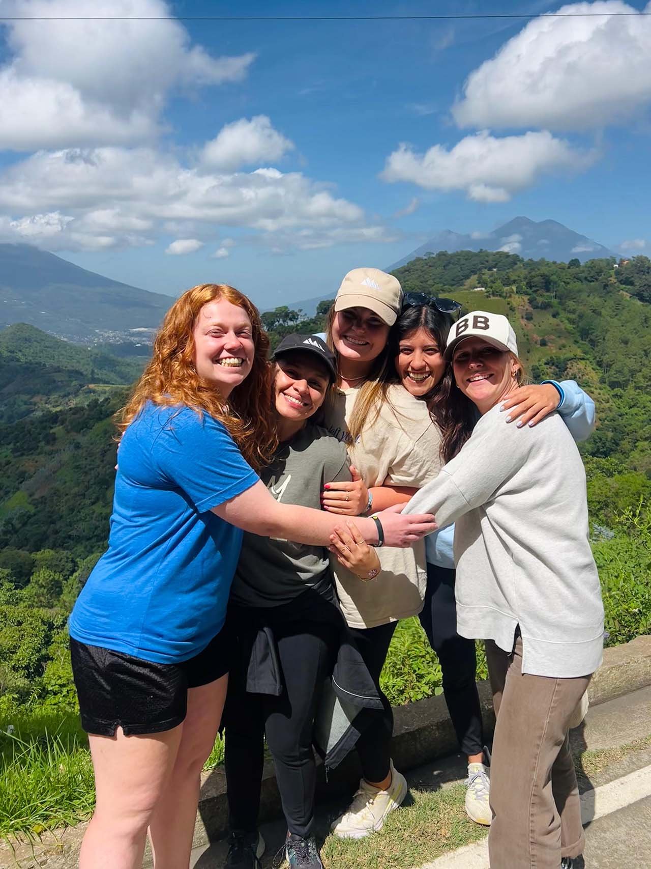 Five women outdoors with mountains and green hills in the background, smiling and hugging each other.