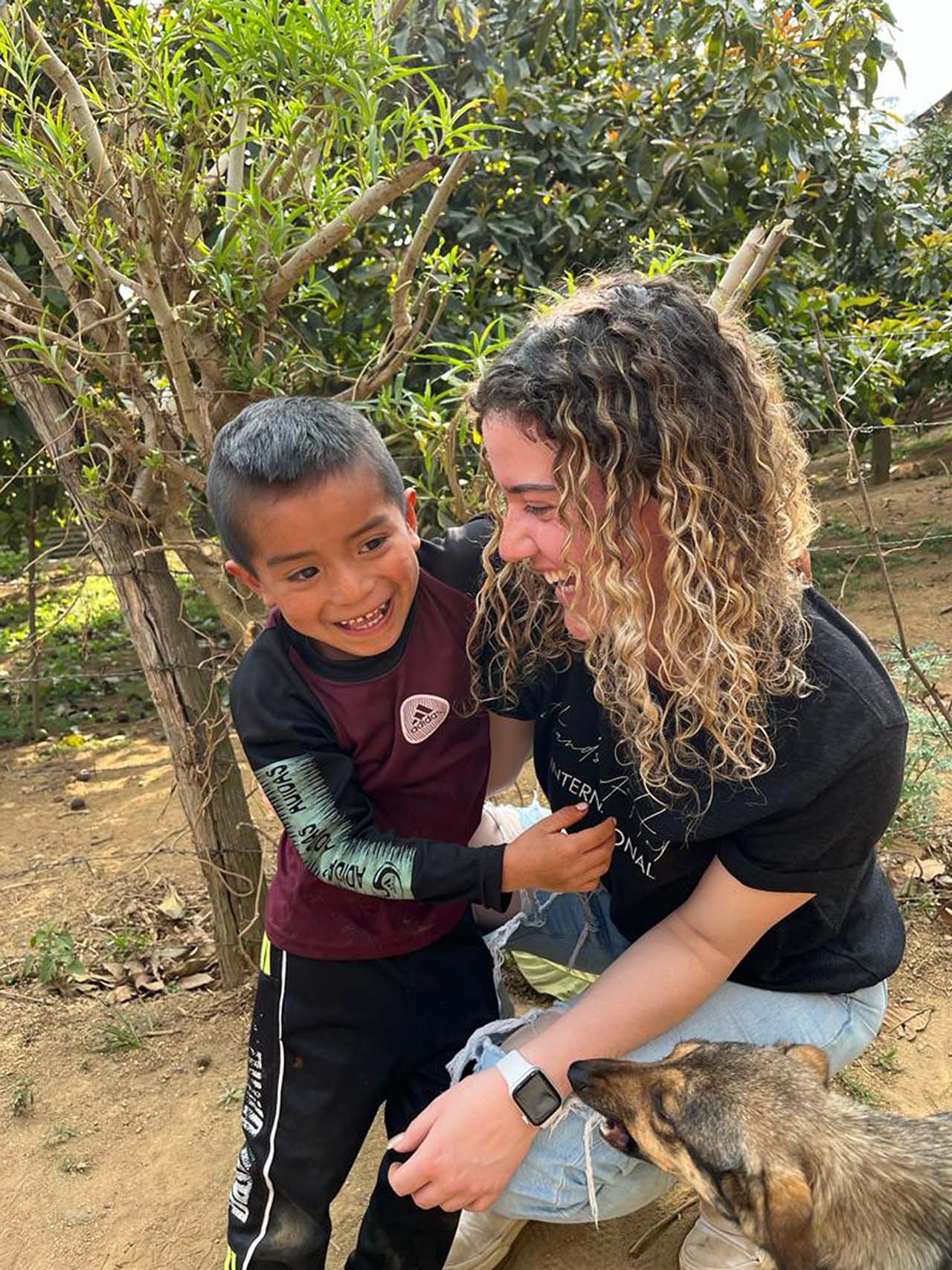 A woman and a young boy are playing together outside, smiling and laughing. A small dog is nearby, looking up at them. The scene is set in a natural environment with trees and bushes.