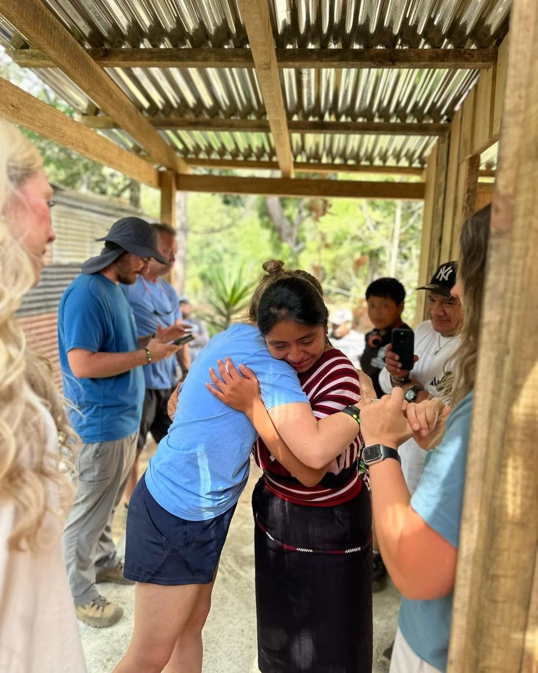A group of people, including a woman in a red and black dress and a woman in a blue shirt, are engaged in a group hug under a wooden shelter with a corrugated metal roof. The background shows trees and outdoor scenery.