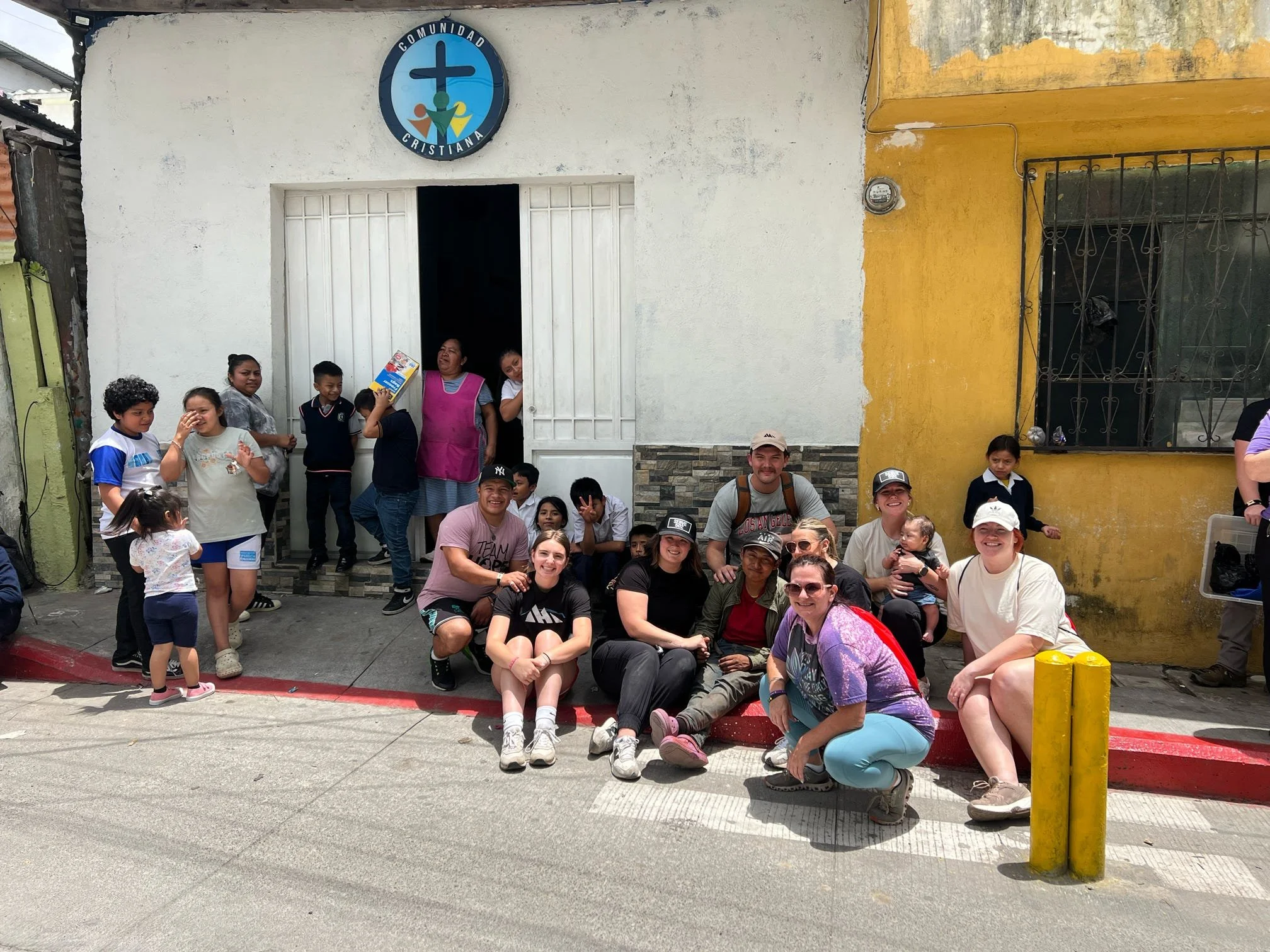 A group of people, including children and adults, gathered outside a community church with a blue and white sign above the entrance that reads "Comunidad Cristiana". Some are sitting on the curb, while others are standing in and near the entrance, posing and smiling for the photo. The background shows a yellow and white building with barred windows.