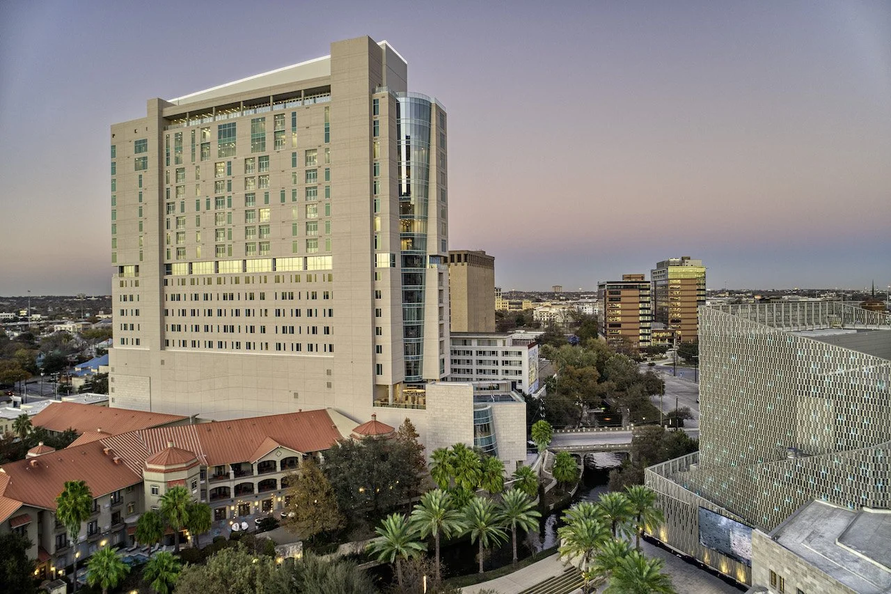 A cityscape at dusk with tall modern buildings, palm trees, and a river running through the scene.
