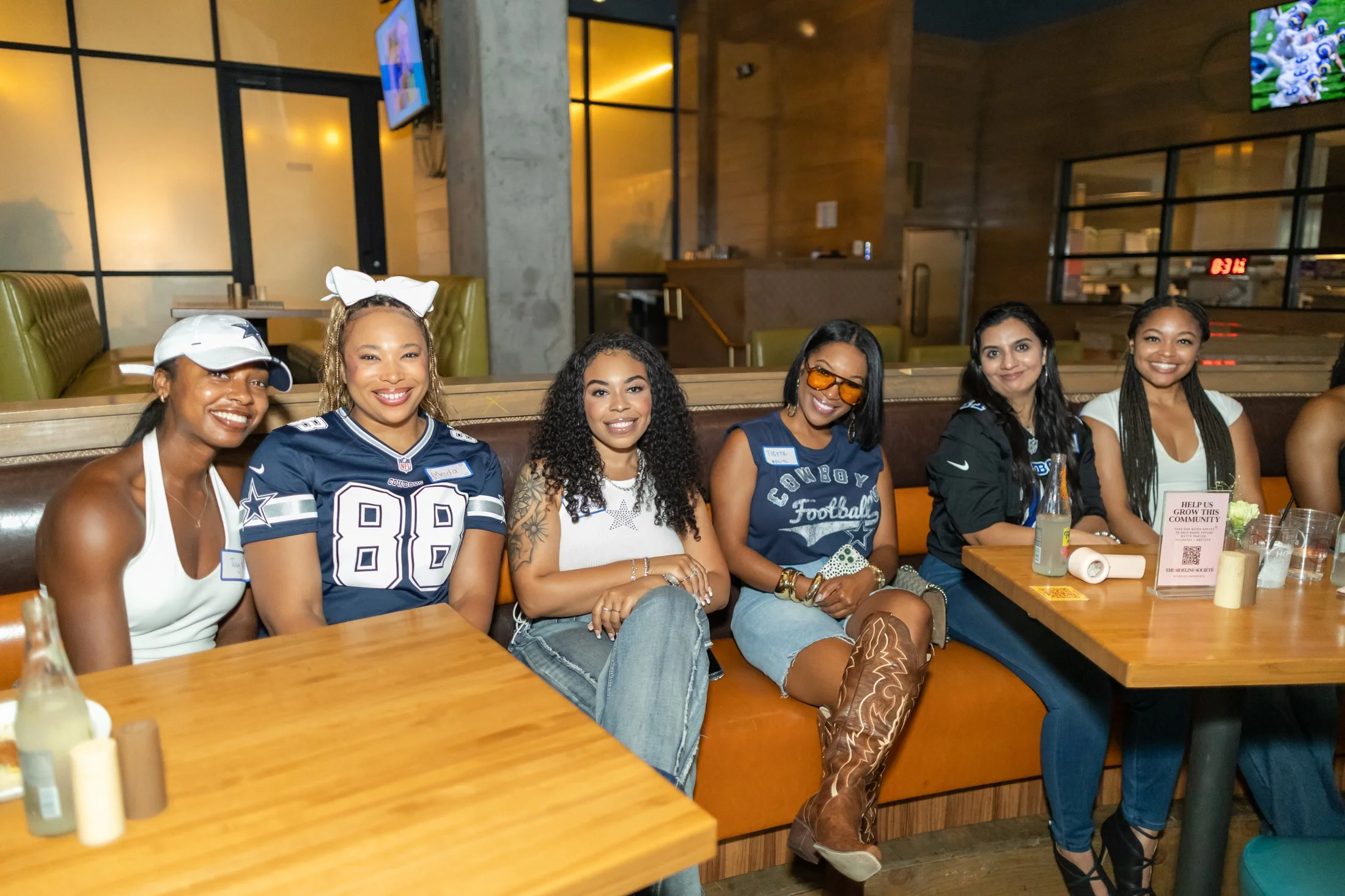 Six women sitting at a restaurant table, smiling and posing for the photo. They are celebrating and dressed in casual and sportswear, with a background of restaurant decor and a TV screen.