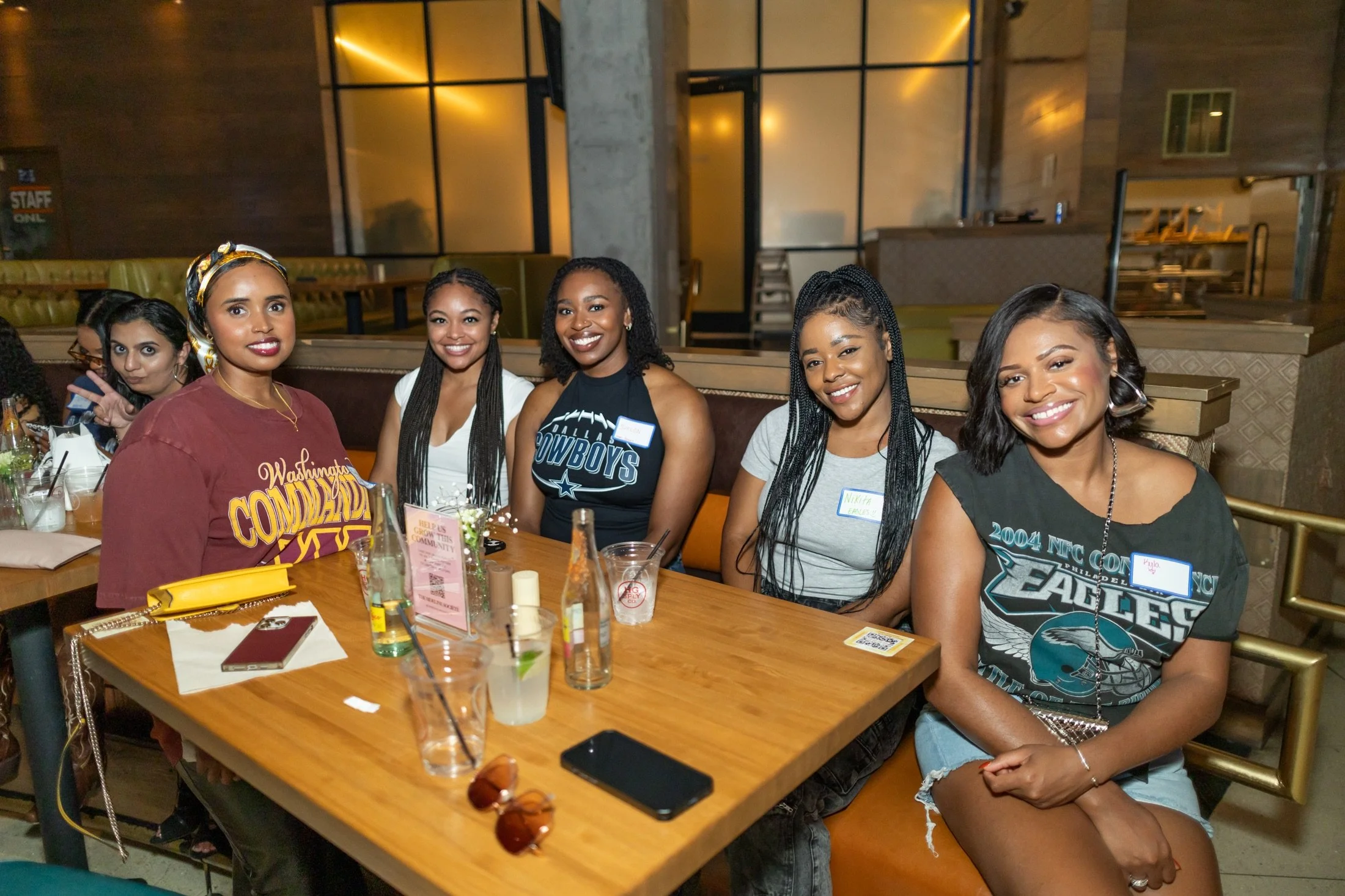 Group of five women sitting at a restaurant table, smiling, with drinks and belongings on the table.