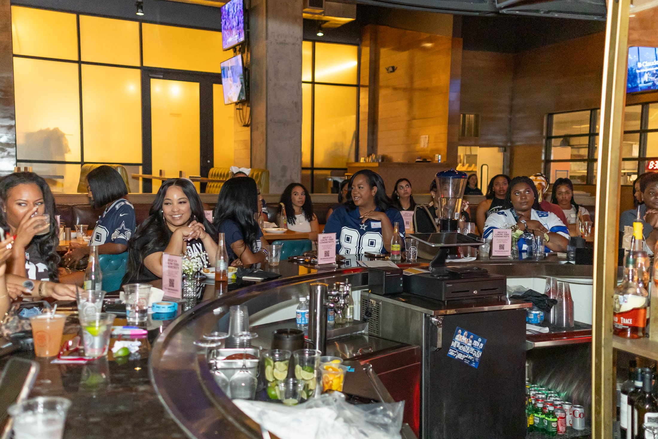 A group of women in sports jerseys sitting at a bar, some laughing and chatting, with drinks and snacks on the counter, in a lively restaurant or bar setting.