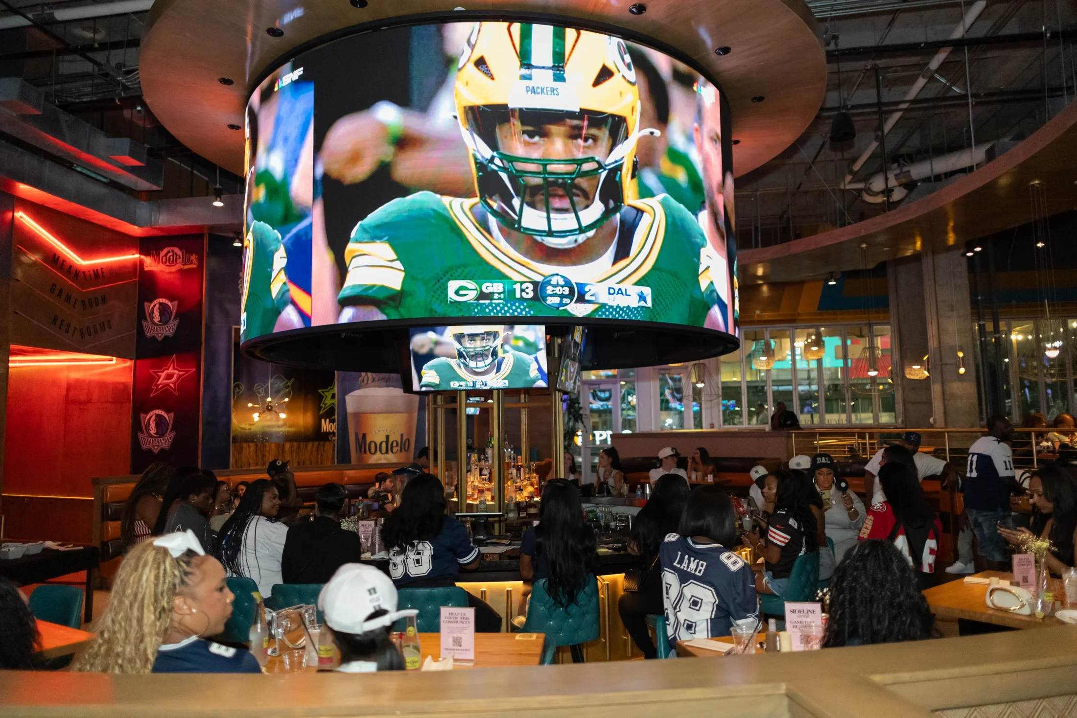 People watching a football game on a large indoor screen in a sports bar or restaurant, many wearing football jerseys, with tables and drinks around.