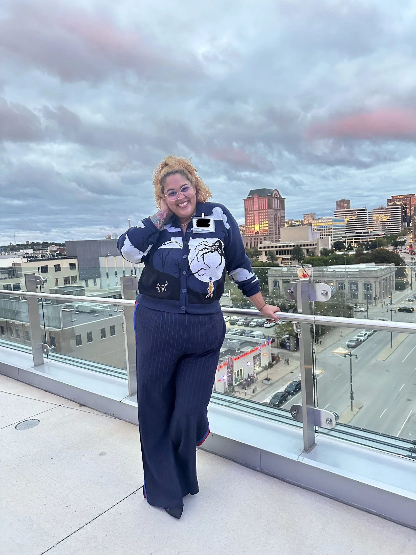 A woman with curly blonde hair, wearing glasses and a navy sweater with white and blue graphic patterns, stands on a rooftop balcony with a city skyline in the background. The sky is cloudy with pinkish hues.