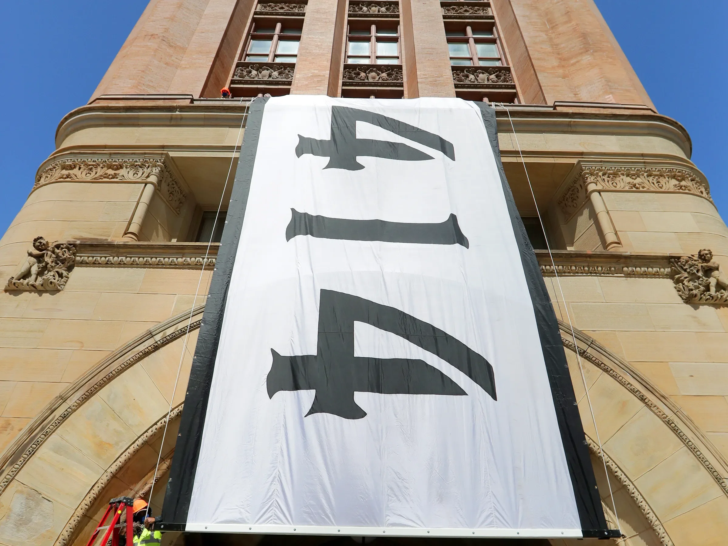 Large white banner with black lettering hanging from a historic brick building, possibly under renovation, with decorative stone carvings and windows visible.