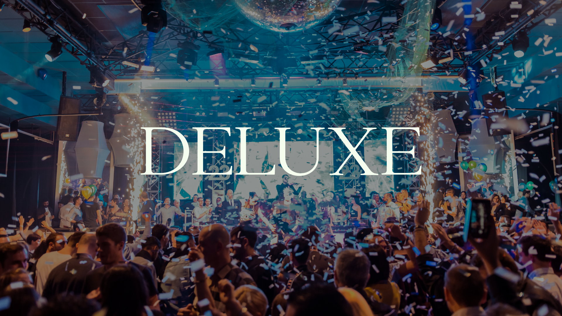 A lively party scene with a large crowd dancing and celebrating in a nightclub or event space with bright lights, confetti, and a DJ on stage, overlaid with the word 'DELUXE' in large white letters.