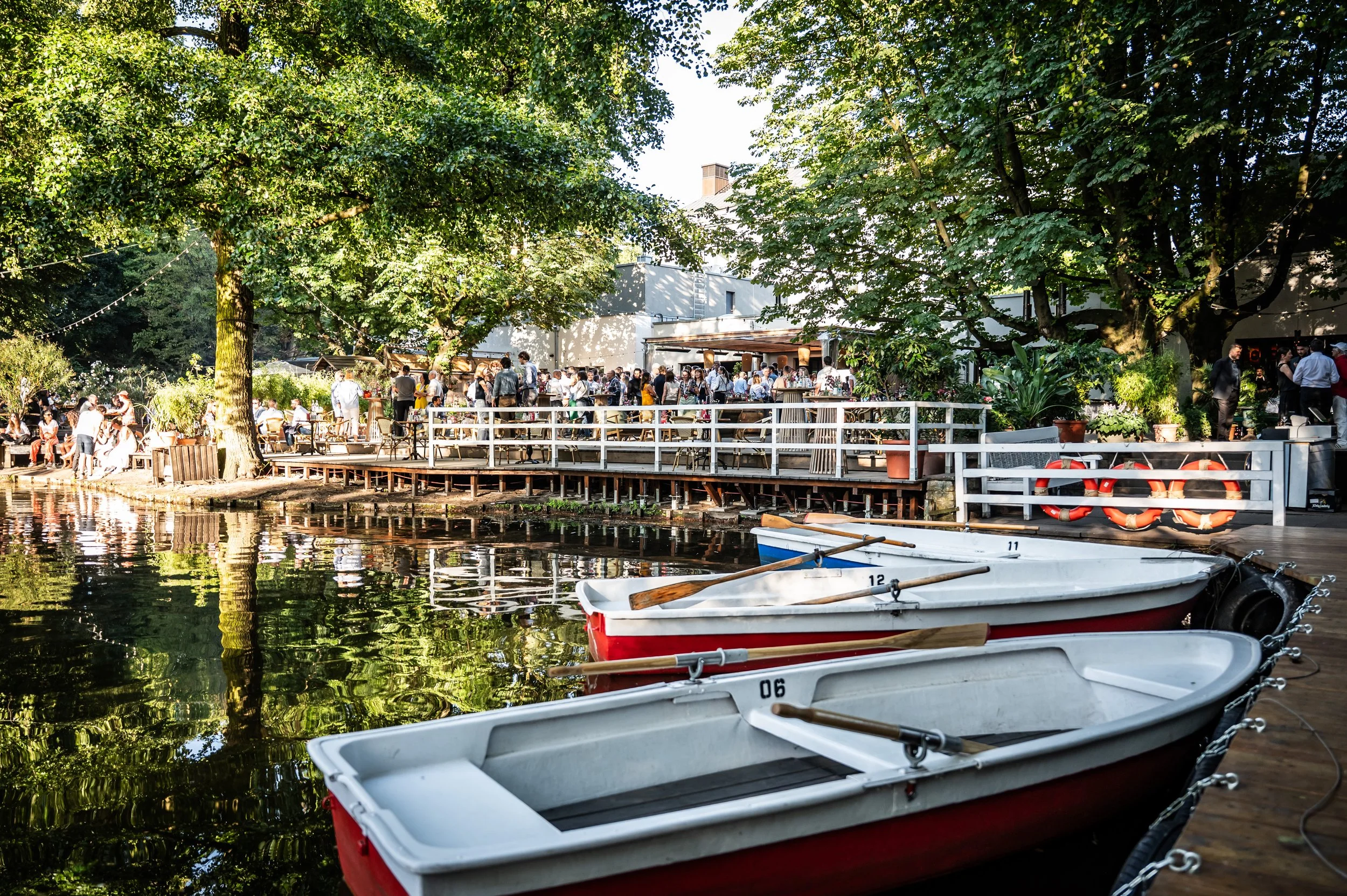 Ein belebter Biergarten am Wasser mit Menschen, die sitzen und sich unterhalten, einige stehen in der Nähe eines Tisches, im Vordergrund sind drei Boote am Ufer festgemacht, im Hintergrund sind große Bäume und ein Gebäude sichtbar.