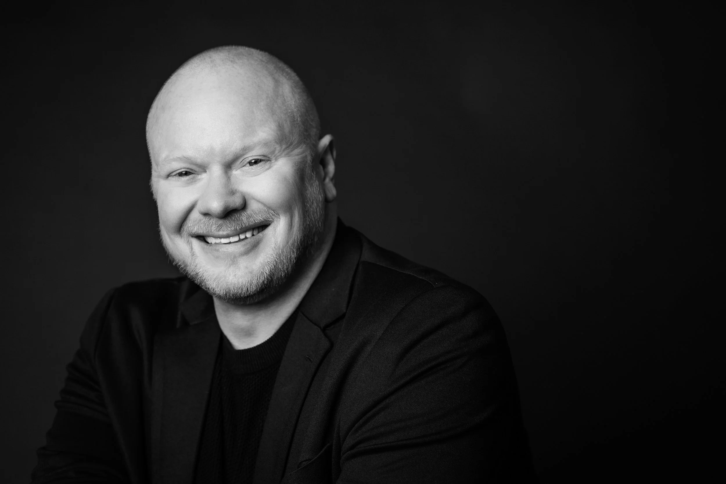 Schwarz-weiß Portrait eines lächelnden Mannes mit kurzen Haaren und Bart, trägt dunkle Kleidung, vor dunklem Hintergrund.
