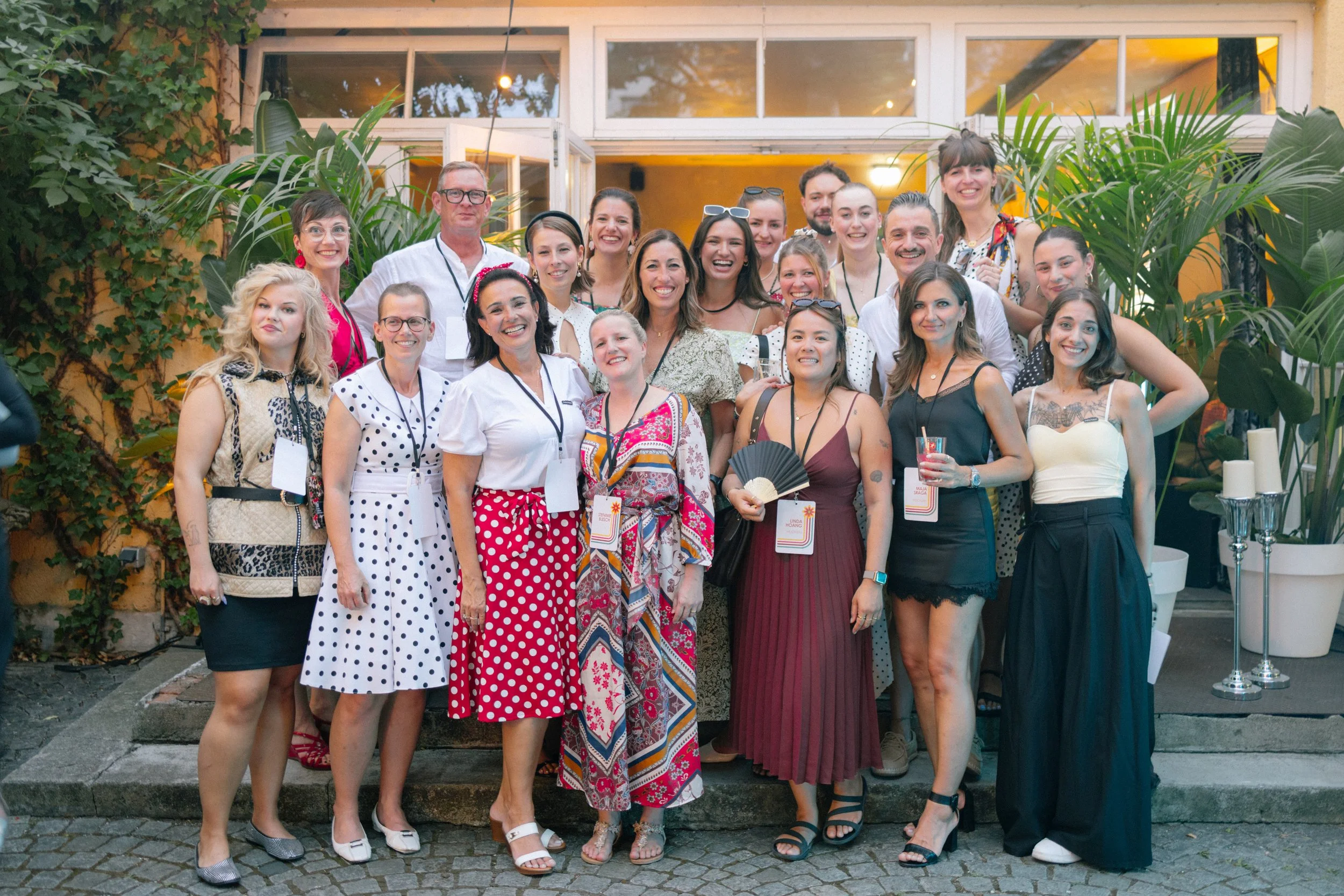 A group of diverse women and men smiling at a social gathering outdoors, standing on steps in front of a building with large windows and surrounded by lush green plants.