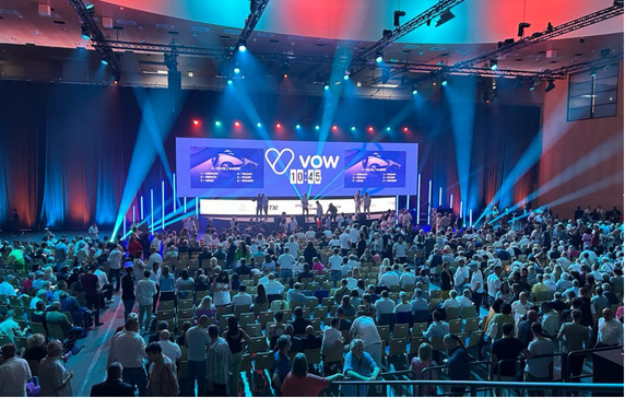 Große Konferenzhalle mit einer Bühne, auf der eine Präsentation stattfindet. Das Publikum sitzt in Reihen, es gibt bunte Bühnenbeleuchtung und einen großen Bildschirm mit dem Logo 'VOW'.