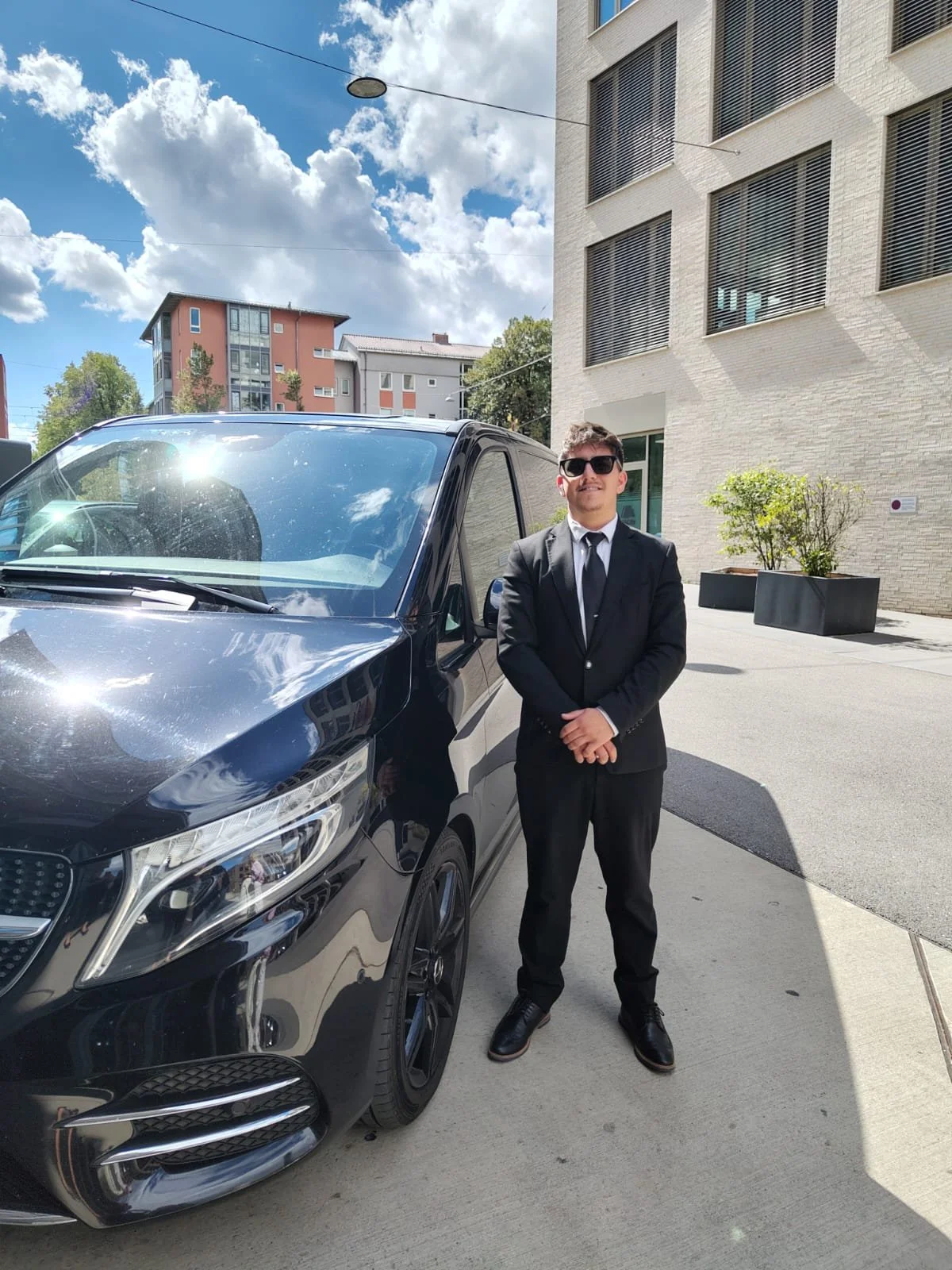 A man in a black suit, white shirt, and black tie standing next to a black car outdoors on a sunny day with partly cloudy sky, modern buildings, and potted plants in the background.