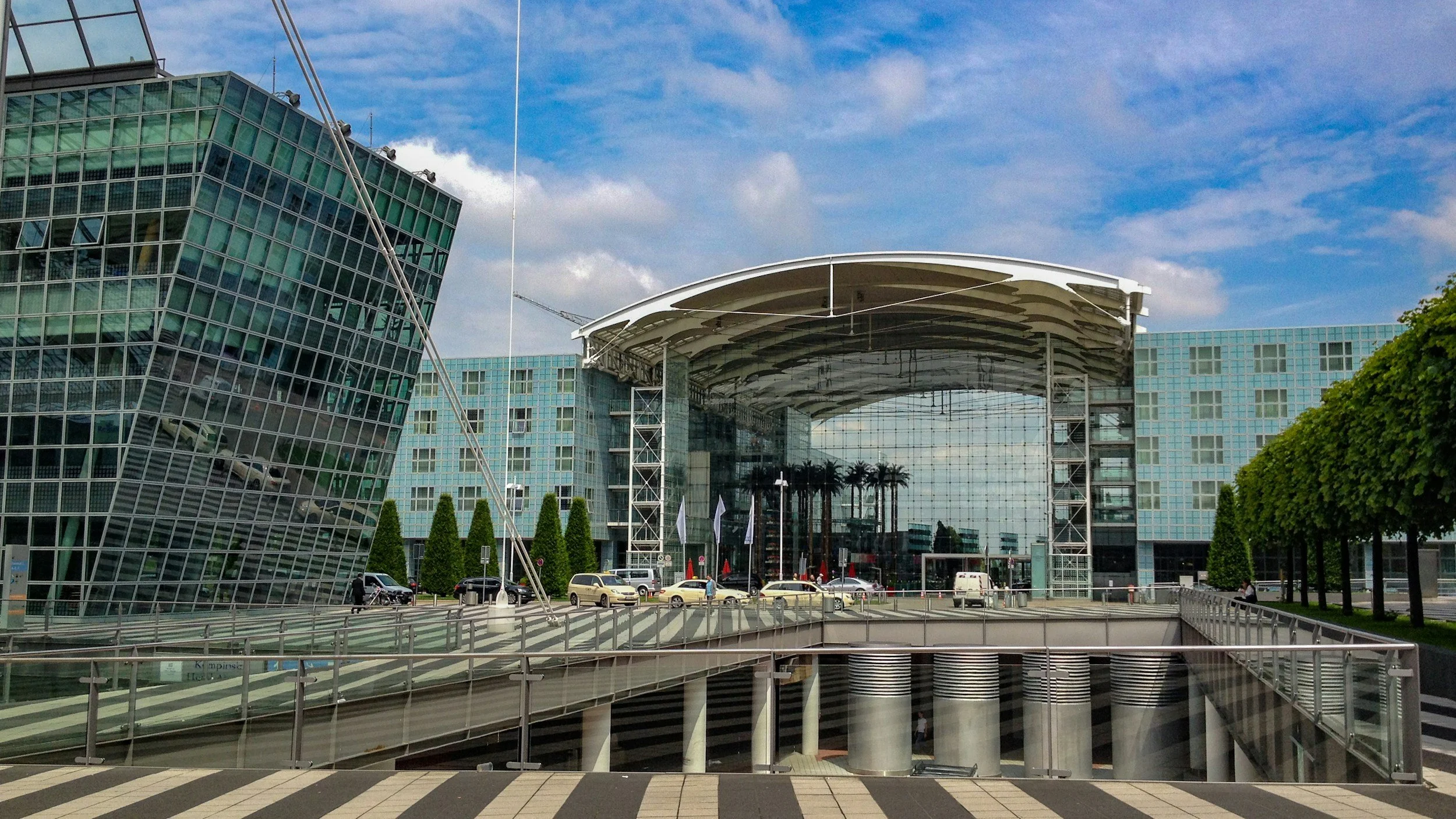 Modernes Bürogebäude mit Glasfassade und offenem Eingangsbereich, Palmen im Hintergrund, Fahrzeuge auf der Straße, Himmel mit Wolken.