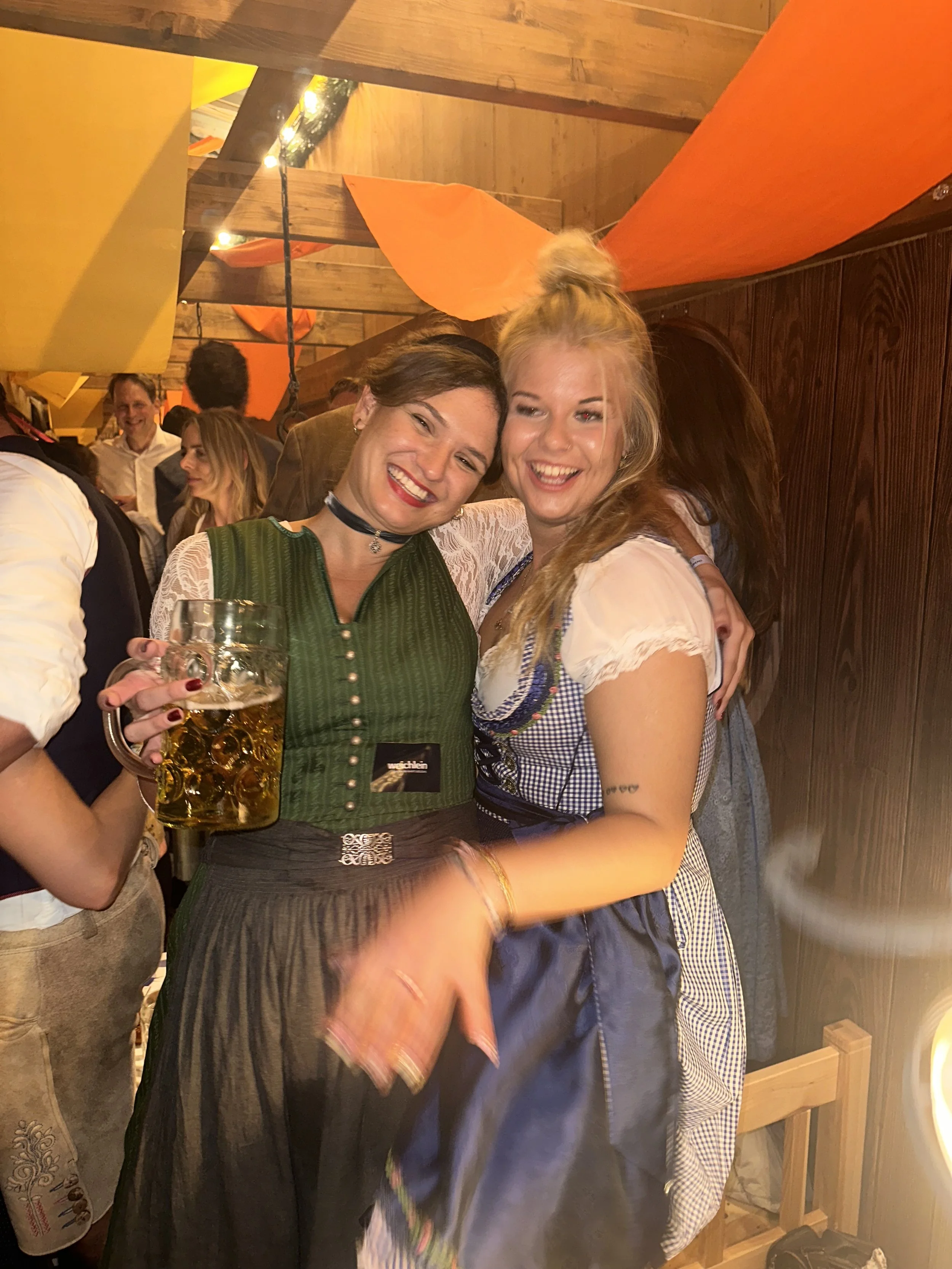 Zwei lachende Frauen in traditionellen Trachten beim Feiern, eine hält ein großes Bier in der Hand, die andere trägt ein weißes Dirndl mit blauer Gürtel, im Hintergrund mehrere weitere Menschen in festlicher Stimmung