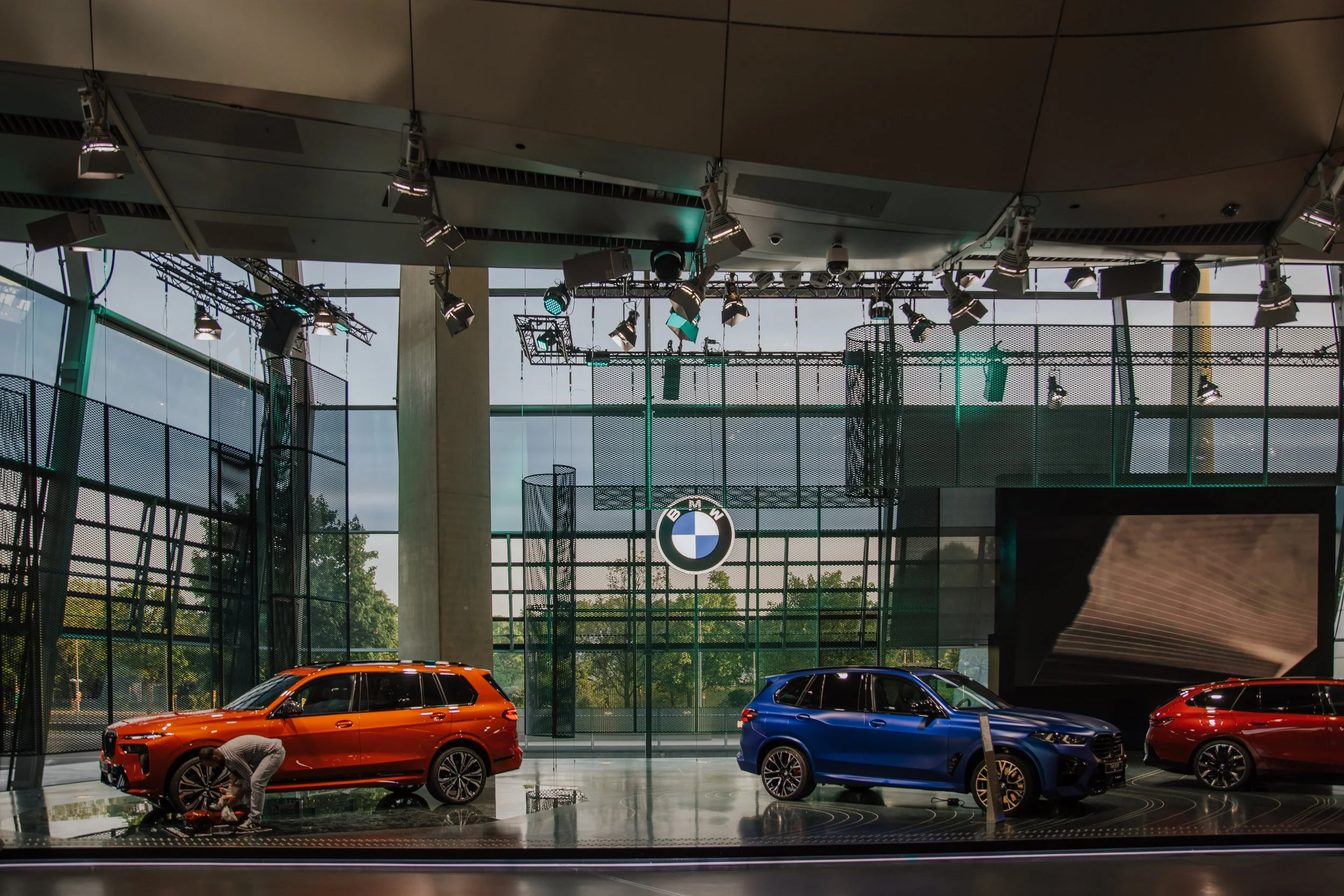Innenansicht einer Autohalle mit drei geparkten Fahrzeugen, einem orangefarbenen SUV, einem blauen SUV und einem roten Kombi, sowie einem Mann, der am orangefarbenen Fahrzeug arbeitet. Die Halle hat große Fenster, eine Decke mit Bühnenbeleuchtung und eine große BMW-Logo auf der Wand im Hintergrund.