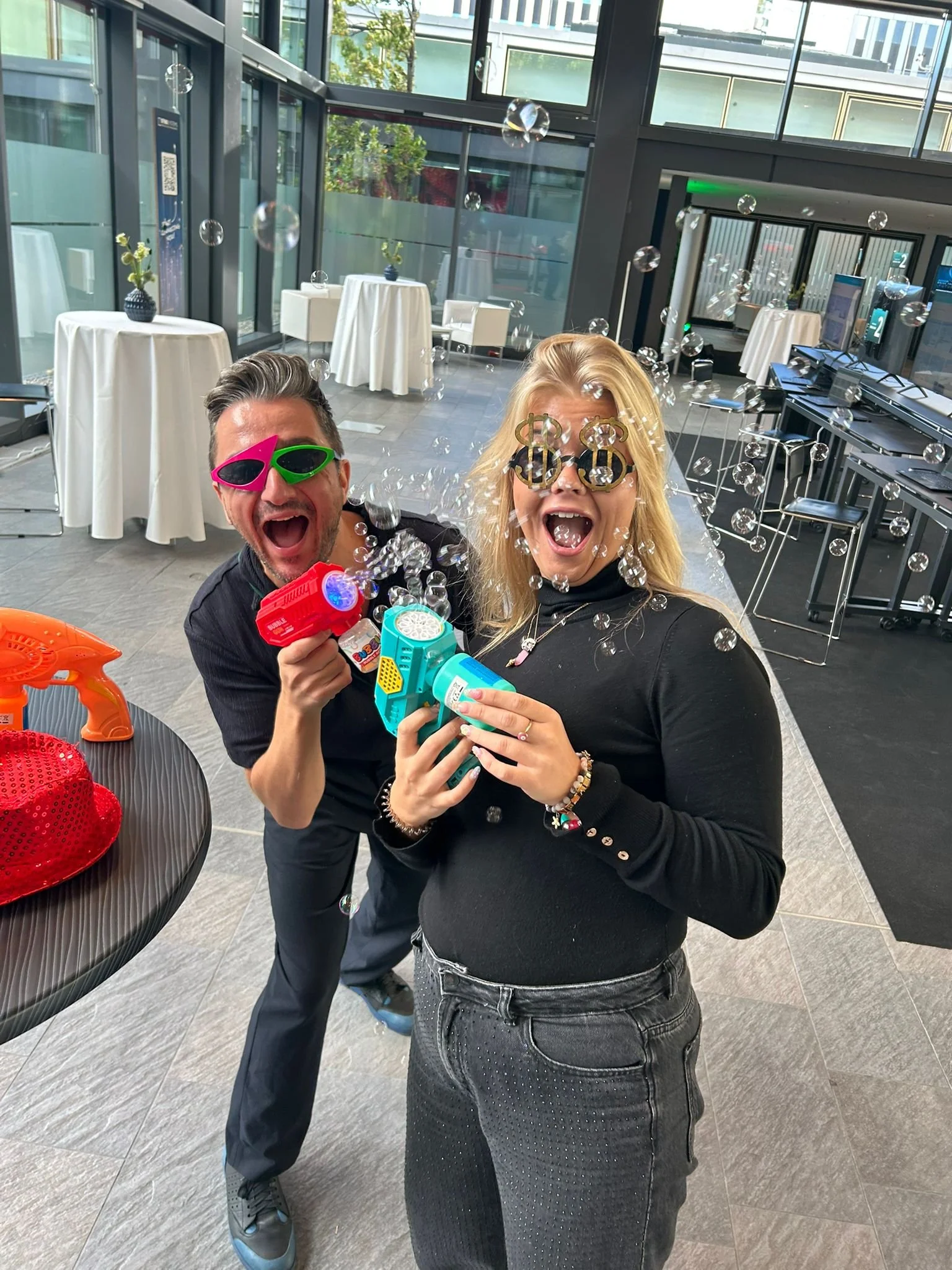 Zwei lachende Menschen in Party-Stimmung, tragen lustige Brillen und spielen mit Wasserpistolen, während sie in einem modernen, hellen Raum feiern.
