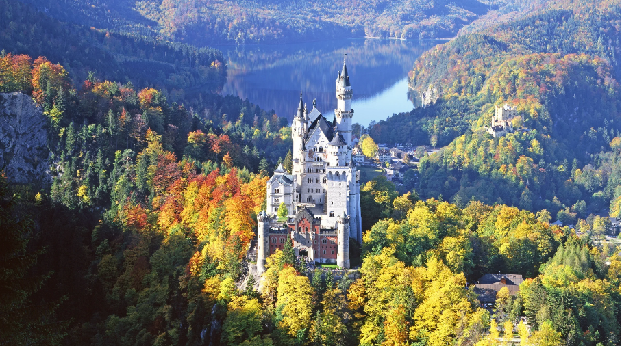 Bayernkönigs Schloss Neuschwanstein auf einem Hügel, umgeben von buntem Herbstlaub, mit einem See im Hintergrund und umgeben von Wäldern.