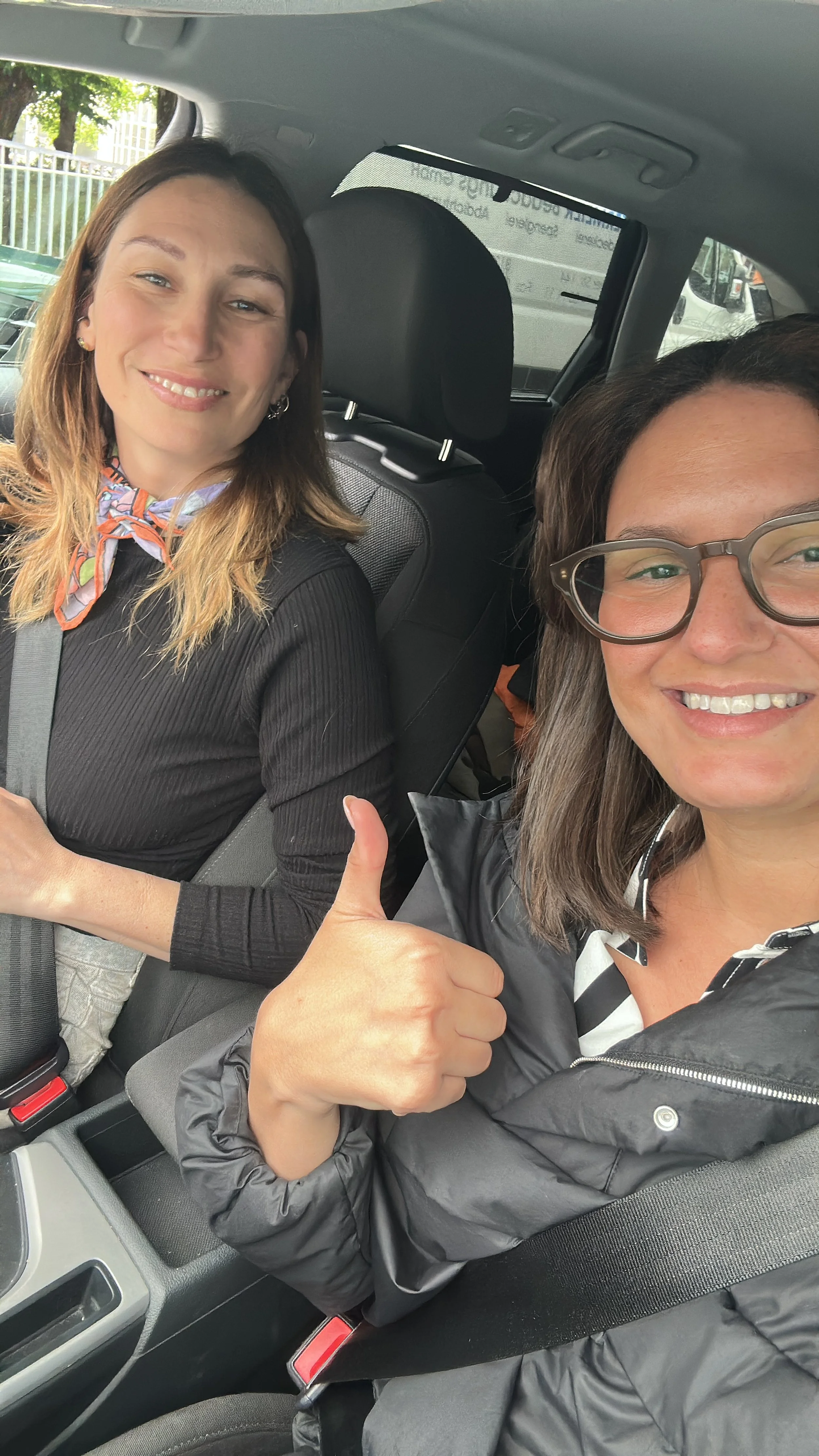 Zwei Frauen sitzen im Auto. Eine Frau trägt eine Brille und zeigt mit dem Daumen nach oben, beide smiling.