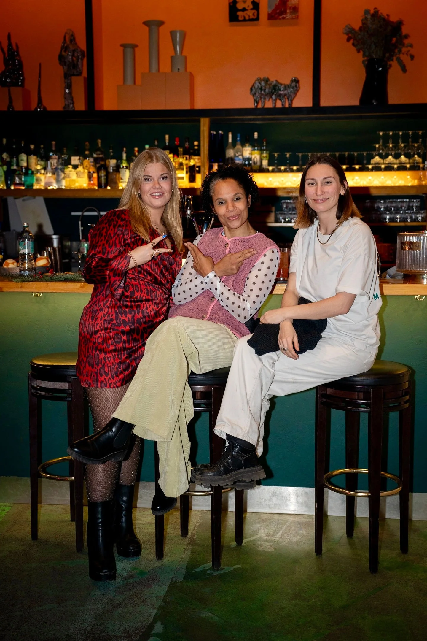 Drei Frauen sitzen an einer Bar, lachen und posieren für die Kamera, im Hintergrund sind Spirituosen und Gläser sichtbar.