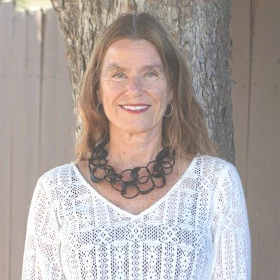 A woman with long brown hair, wearing a white lace top and a statement black necklace, standing outdoors in front of a tree and wooden fence, smiling at the camera.