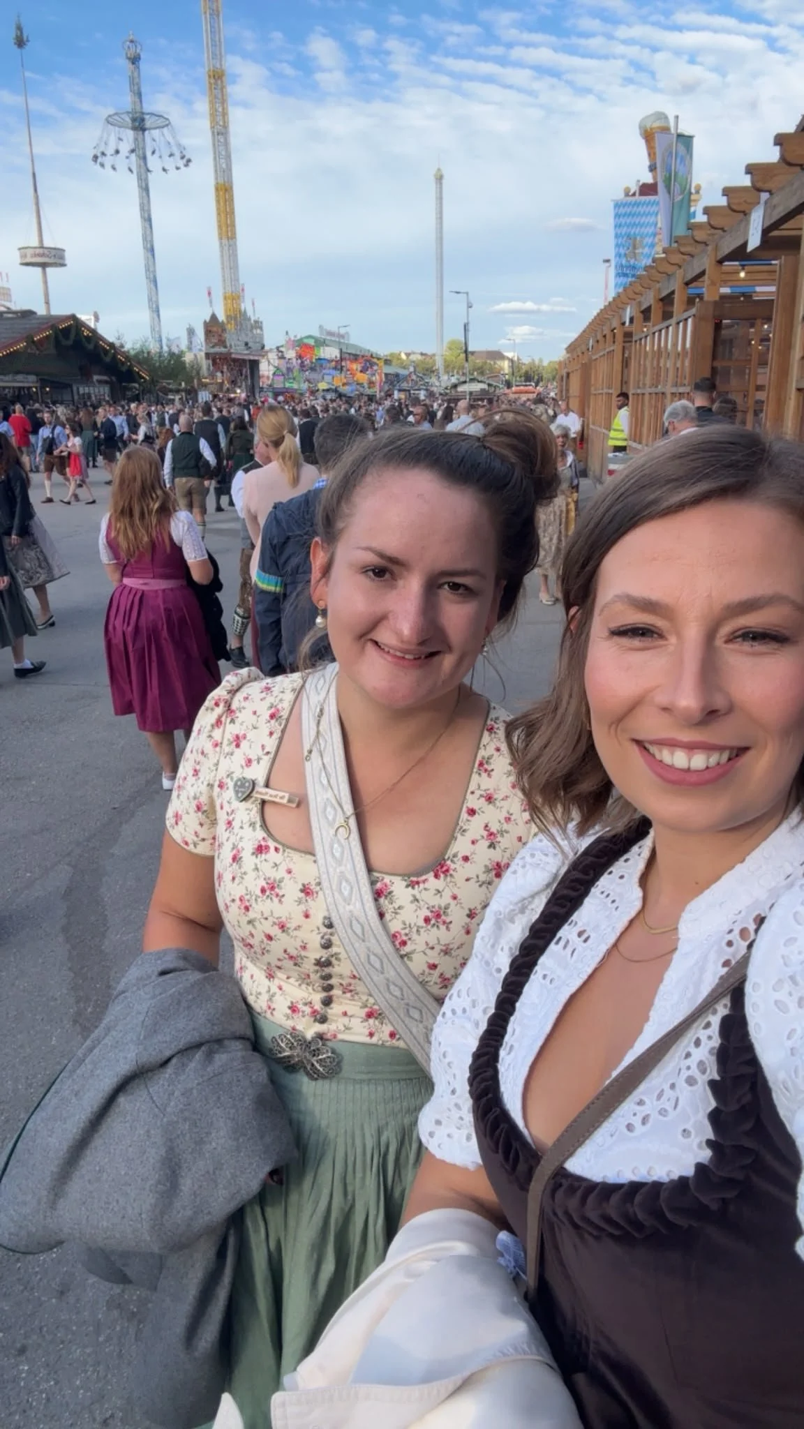 Two young women taking a selfie at a crowded amusement park with rides and attractions in the background.