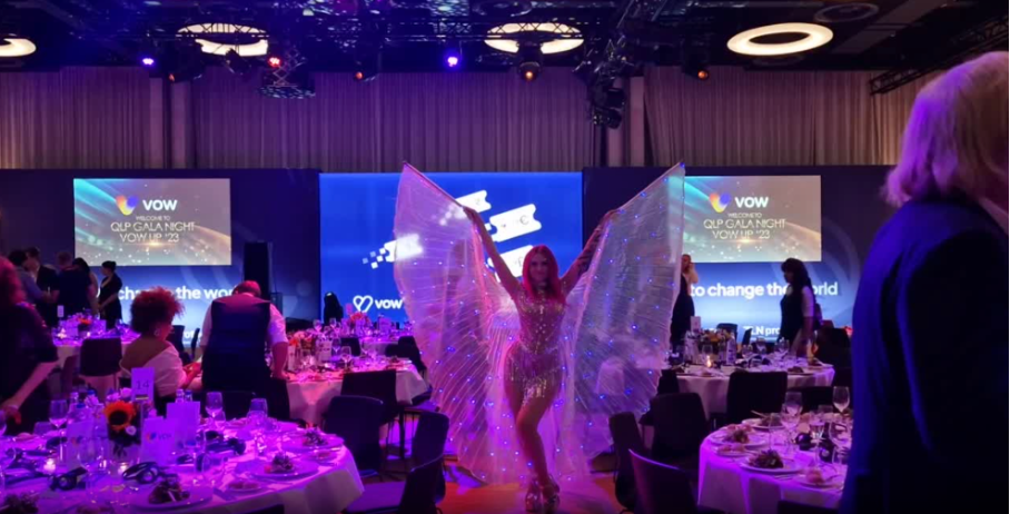 Frau in einem glitzernden Kostüm mit großen, leuchtenden Flügeln tanzt in der Mitte eines festlich dekorierten Saals während einer Gala-Veranstaltung.