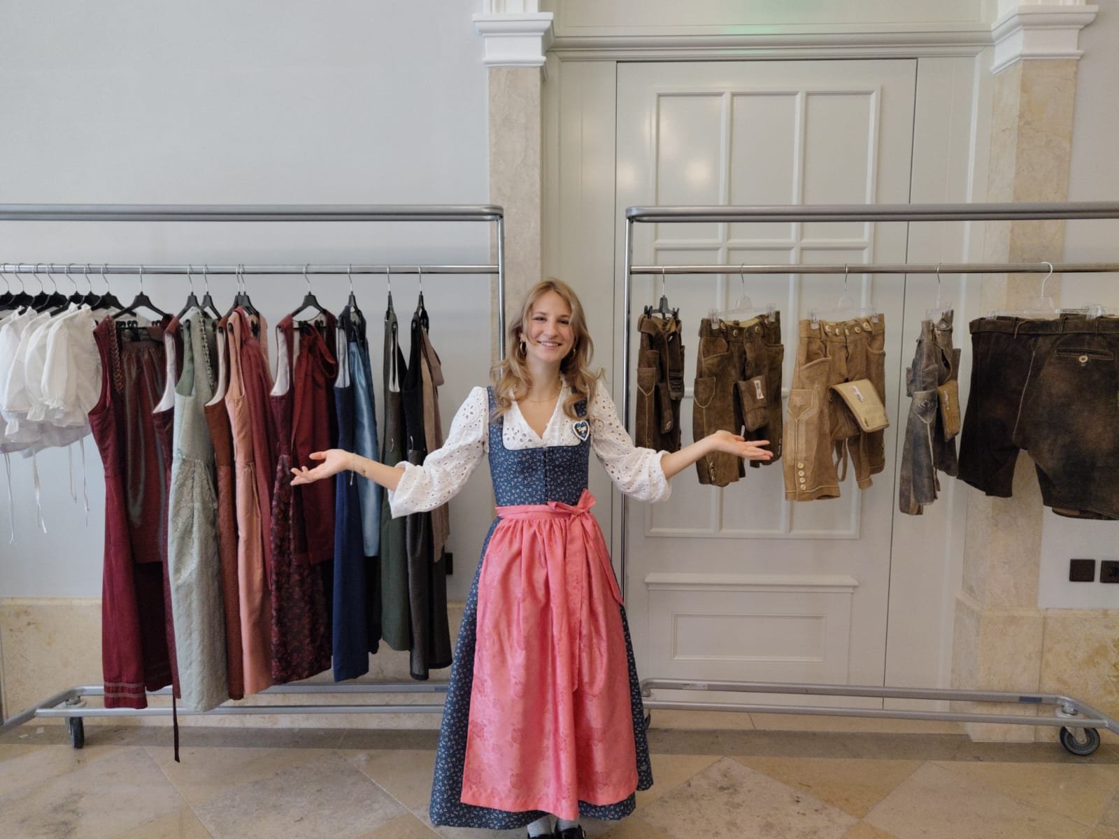 A woman dressed in traditional German dirndl standing in a clothing store or exhibition, with racks of dresses and lederhosen behind her.