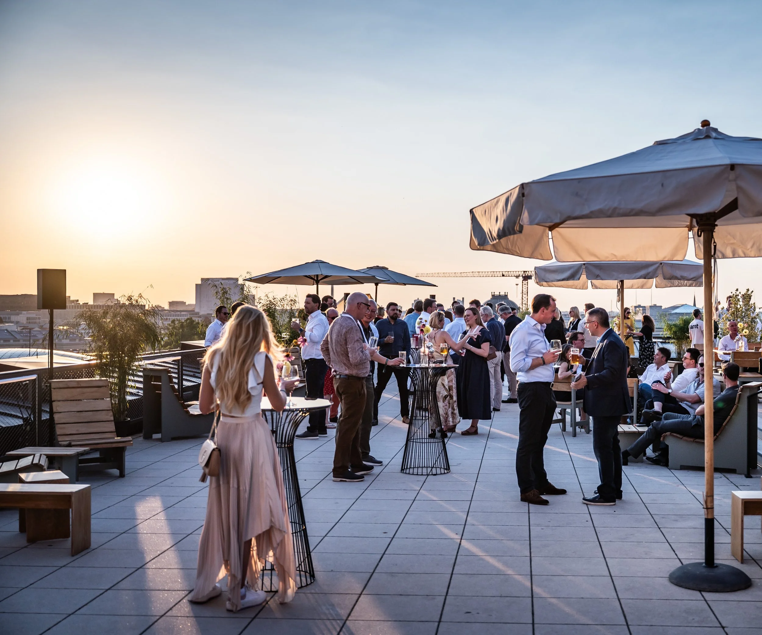 Menschen auf einer Dachterrasse bei Sonnenuntergang bei einer ungezwungenen Party mit Getränken, Sonnenschirmen und Blick auf die Stadt