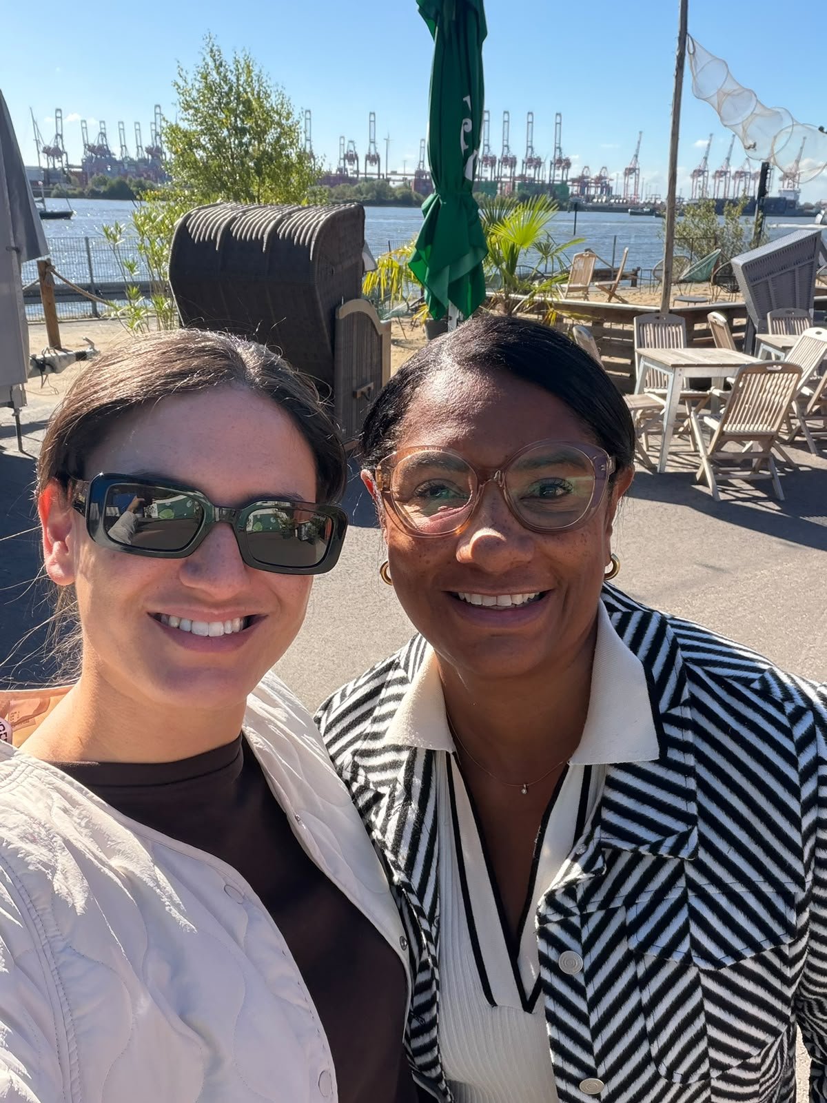 Two women smiling and taking a selfie outdoors by a body of water with docked cranes in the background on a sunny day.