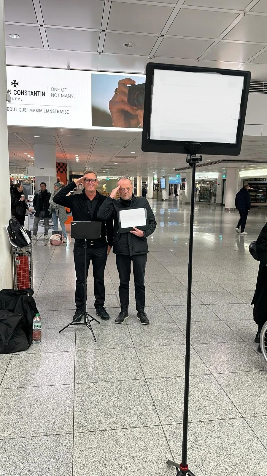 Two men stand in an airport terminal, saluting. One holds a sign and a tablet, while the other holds a sign. There is a large display screen and other travelers in the background.