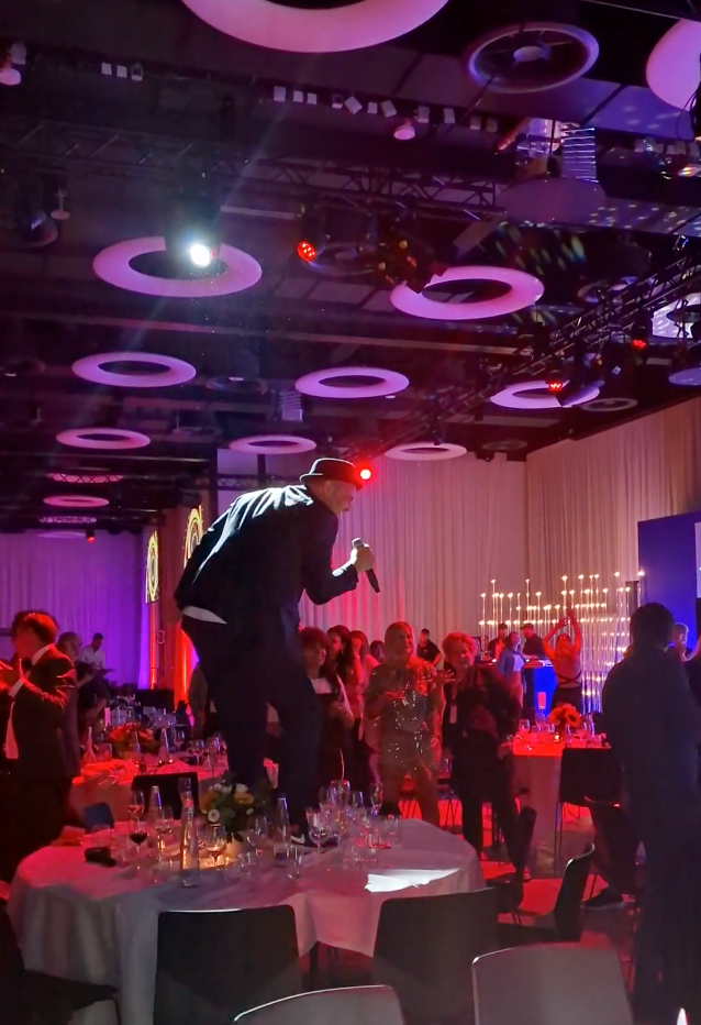 Ein Mann steht auf einem Tisch auf einem Tanzballsaal, hält ein Mikrofon und trägt einen schwarzen Anzug mit Hut. Im Hintergrund tanzen und feiern Menschen bei bunter Beleuchtung und Kerzen. Der Saal ist modern mit kreisförmigen Lichtelementen an der Decke.