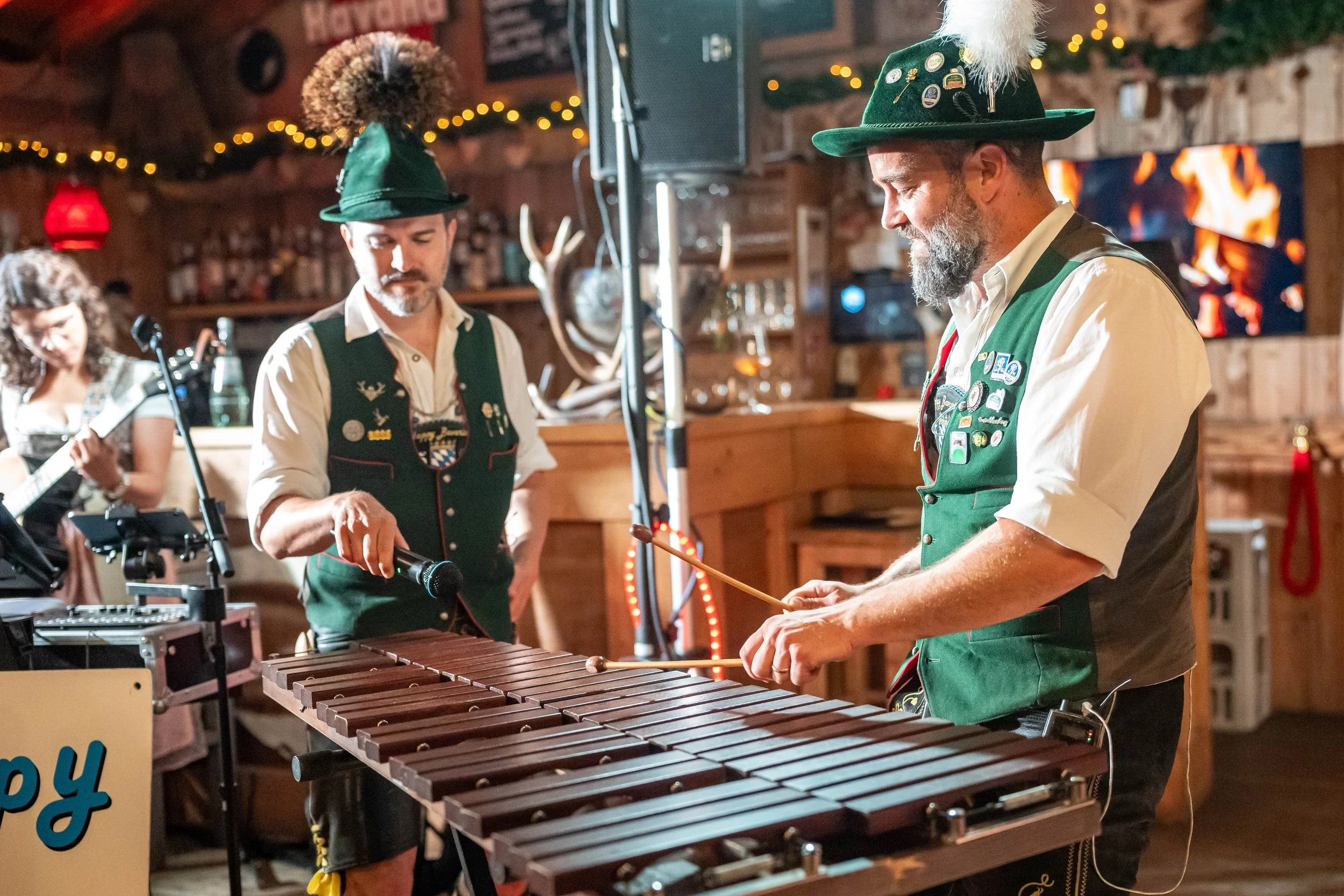 Mehrere Männer in traditionellen Trachten spielen Musikinstrumente, darunter eine Holzblockklaviertafel, in einer festlich dekorierten, rustikalen Umgebung mit Lichterketten.
