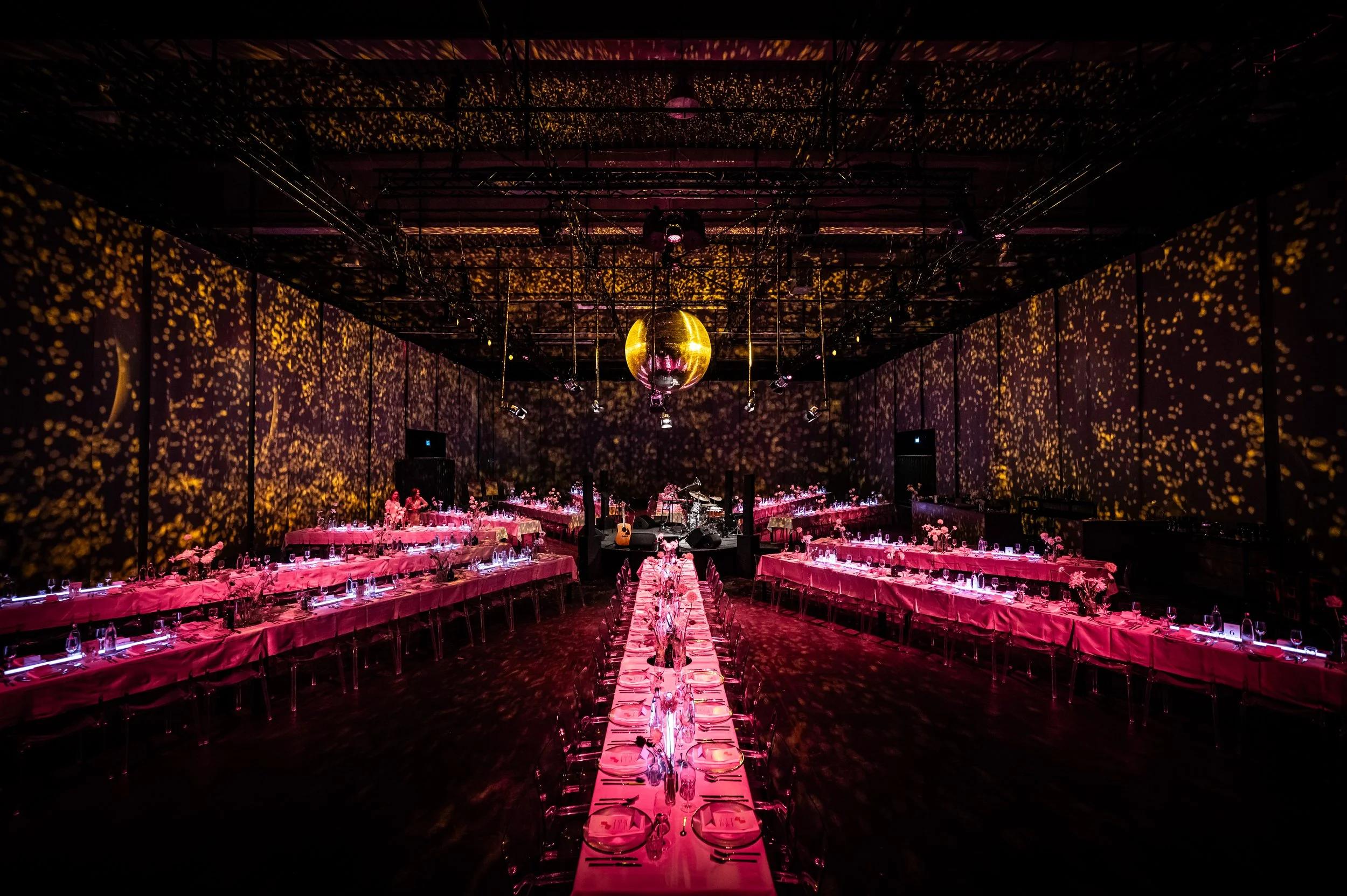 Eleganter Saal mit langen, pinkfarbenen Tischen, einer diskokugel an der Decke und warmer Beleuchtung, die den Raum in goldene Muster taucht.