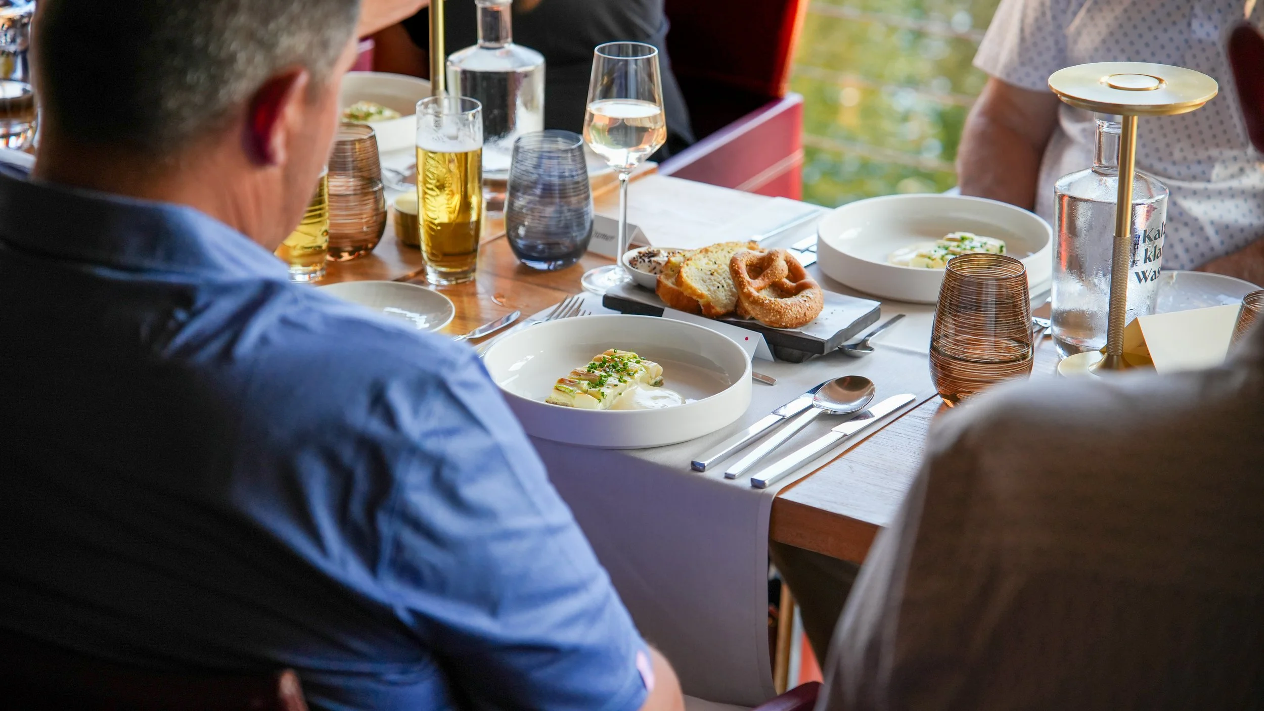 Ein gedeckter Tisch beim Essen mit verschiedenen Tellern, Gläsern und Getränken, im Hintergrund sind mehrere Personen zu sehen.