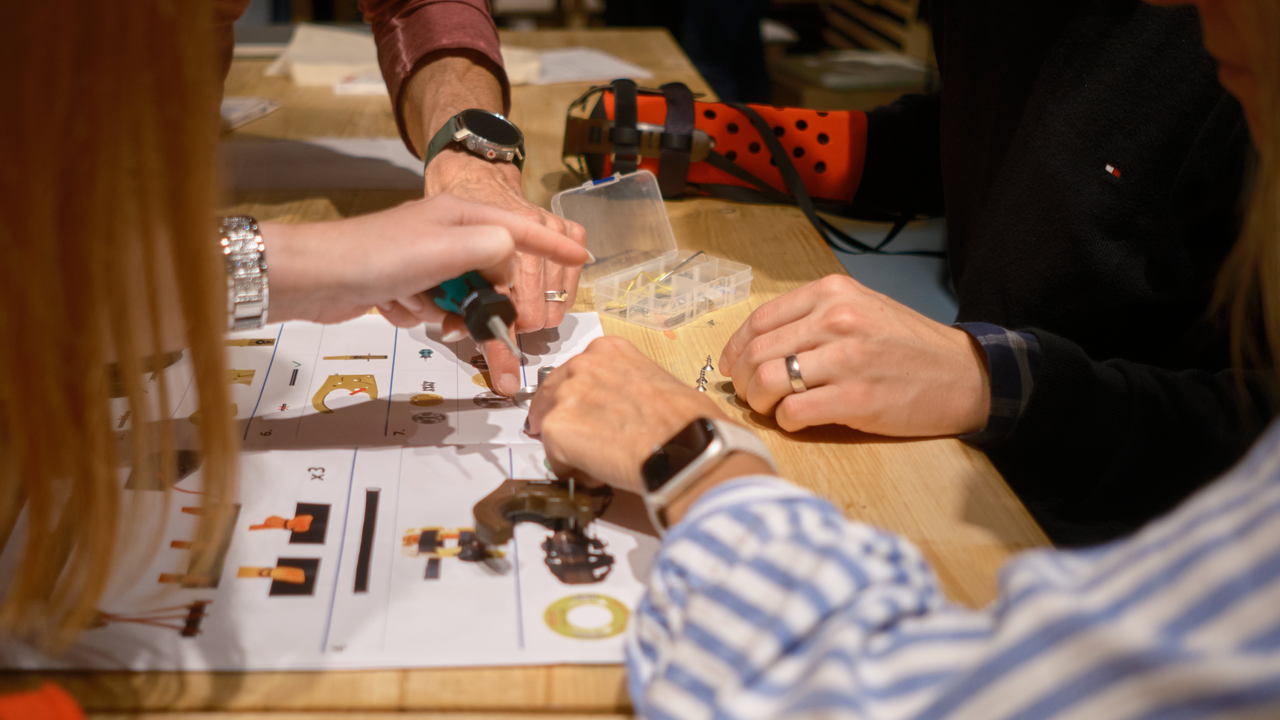 Mehrere Personen bauen mit Werkzeugen und kleinen Teilen an einem Holztisch, Anleitungen und Werkzeugkästen sind sichtbar.