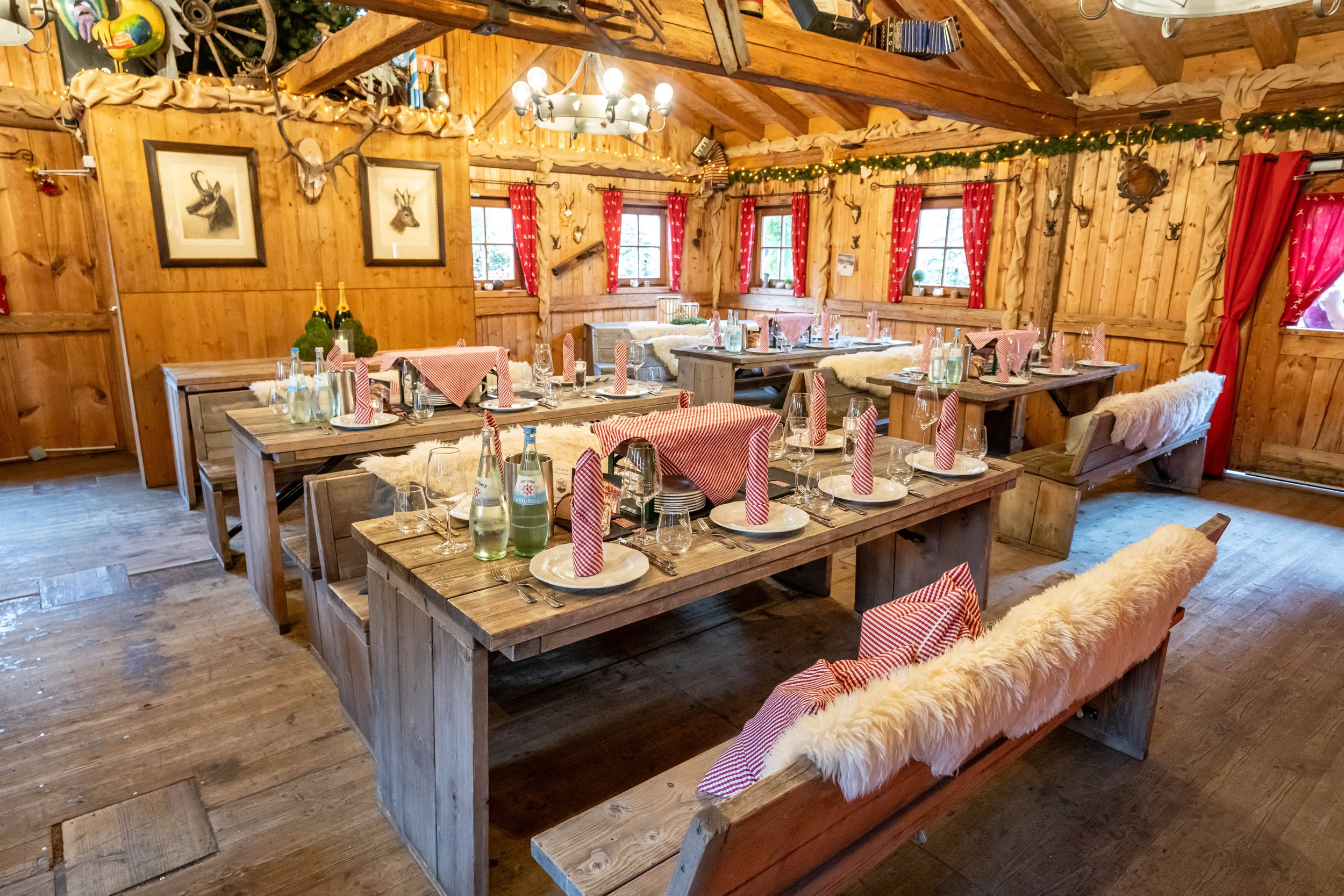 Ein rustikaler, festlich gedeckter Speiseraum im Chalet-Stil mit Holzwänden, -decken und -möbeln. Es sind Tische mit roten und weißen Tischdecken, Wein- und Wasserflaschen sowie Gläser. Die Fenster sind mit roten Vorhängen und Dekorationen im Chalet-Stil geschmückt. Die Beleuchtung erfolgt durch Laternen und eine zentrale Deckenleuchte.