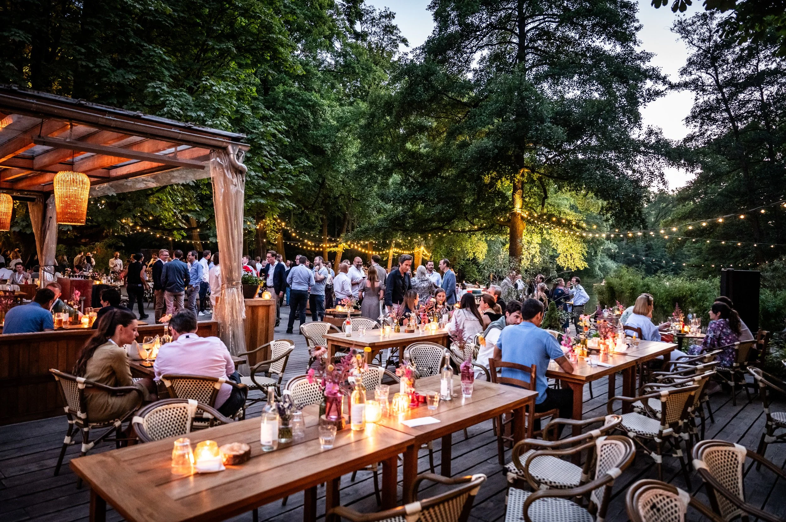 Ebene outdoor Abendveranstaltung mit Tischen, Kerzen, Blumen und vielen Gästen, umgeben von Bäumen, unter Lichterketten