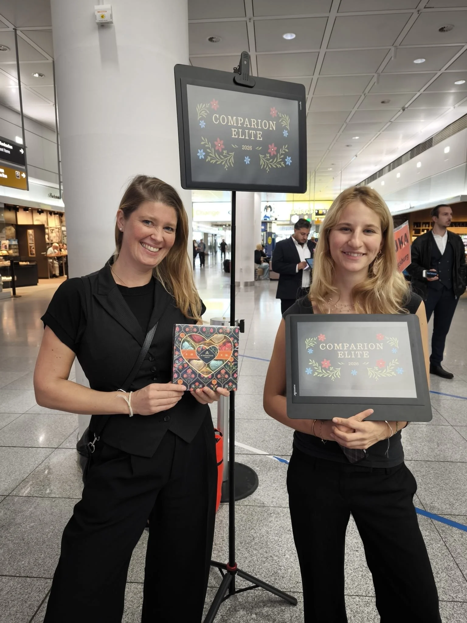 Two women smiling at an airport, holding a sign that says 'Comparison Elite 2026' with floral decorations, standing in front of a digital sign and a group of people in the background.