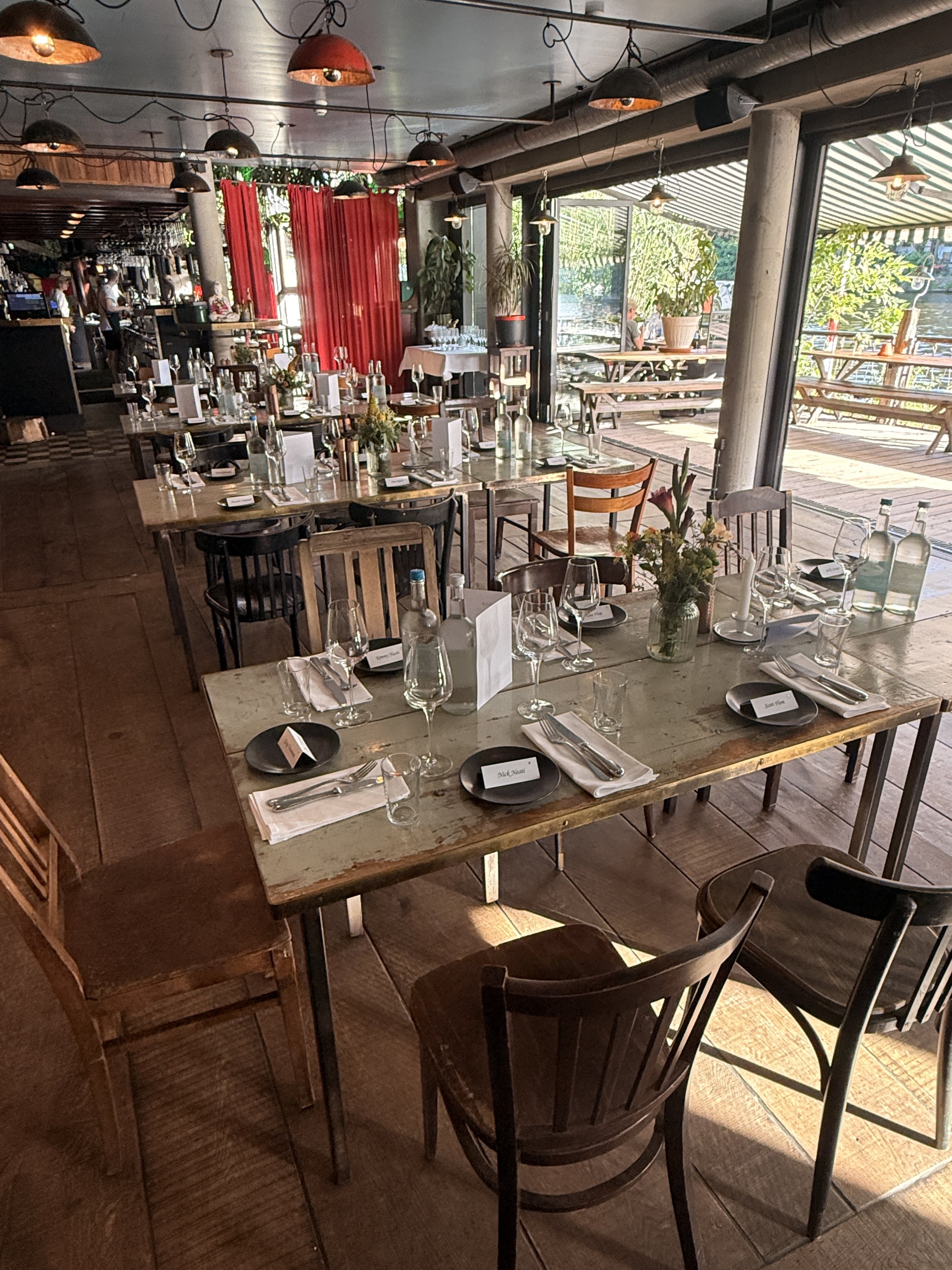 Innenaufnahme eines an einem Wasserweg gelegenen Restaurants mit mehreren Tischen, die für eine Veranstaltung eingedeckt sind, inklusive Gläser, Besteck und Blumen. Außenbereich mit Holztischen ist sichtbar, mit viel Sonnenlicht und grünen Pflanzen.