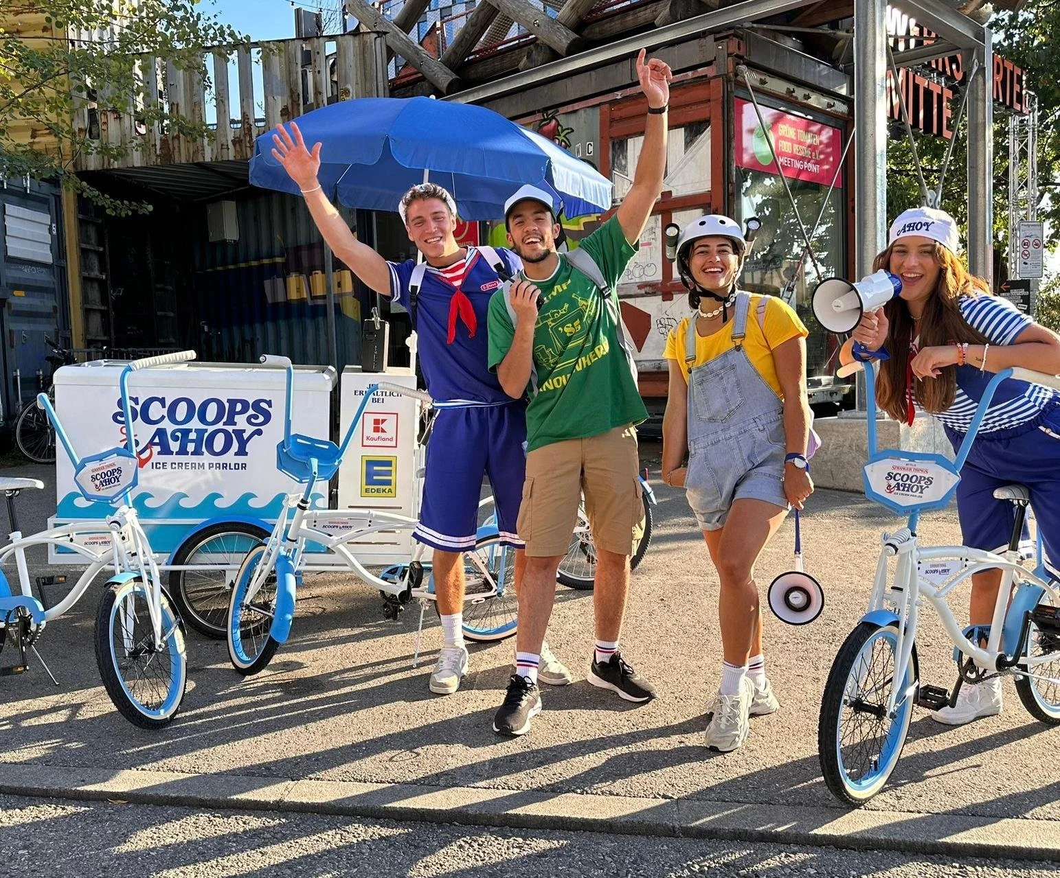 Vier junge Menschen stehen lachend neben weißen Fahrrädern mit der Aufschrift "SCOOPS AHOY" vor einem Imbisswagen. Zwei tragen Helme, einer hält ein Megafon, alle freuen sich, bei schönem Wetter.